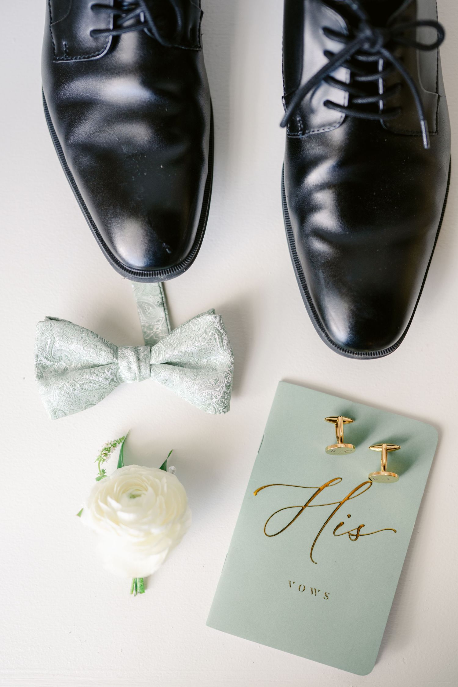 Groom's black dress shoes with mint green bow tie, white boutonniere and vows book arranged on white surface.