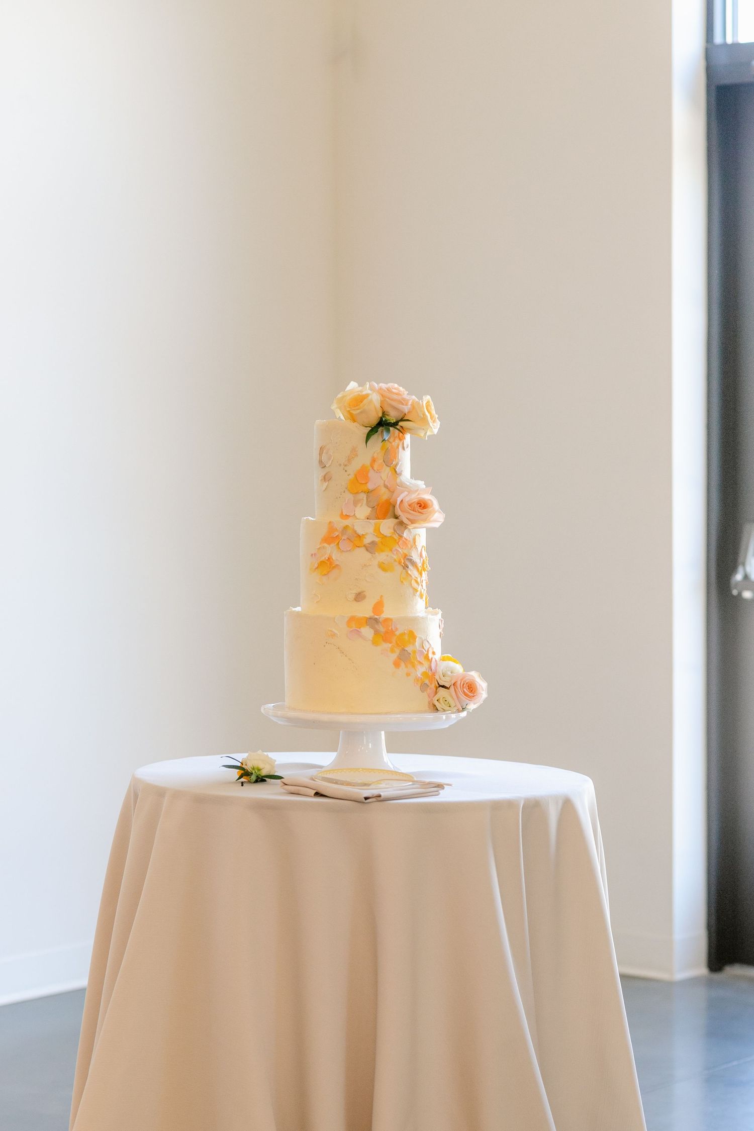 Elegant white wedding cake with peach floral decorations displayed on round table with white tablecloth in bright venue.