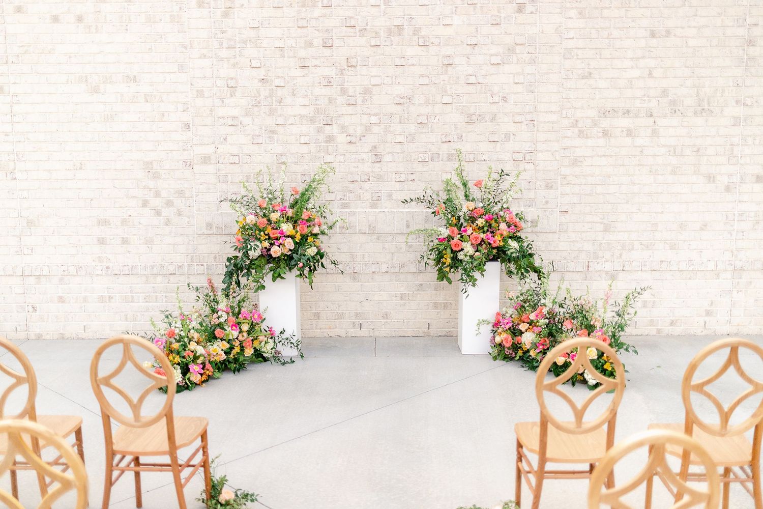 Elegant wedding ceremony setup with white chairs and floral arrangements against a textured stone wall.