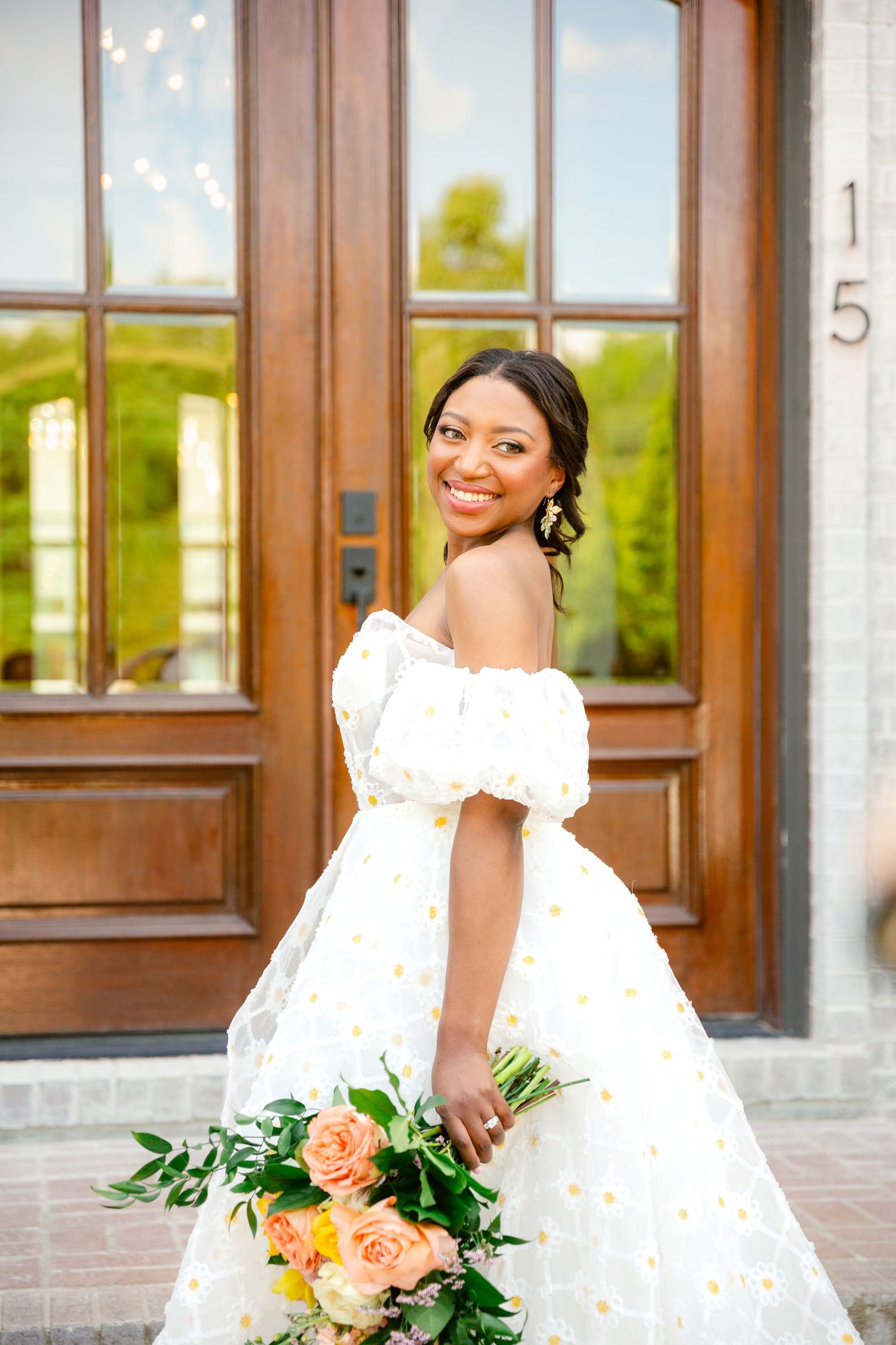 Bride in elegant off-shoulder white wedding dress poses with peach and green floral bouquet near wooden doors.