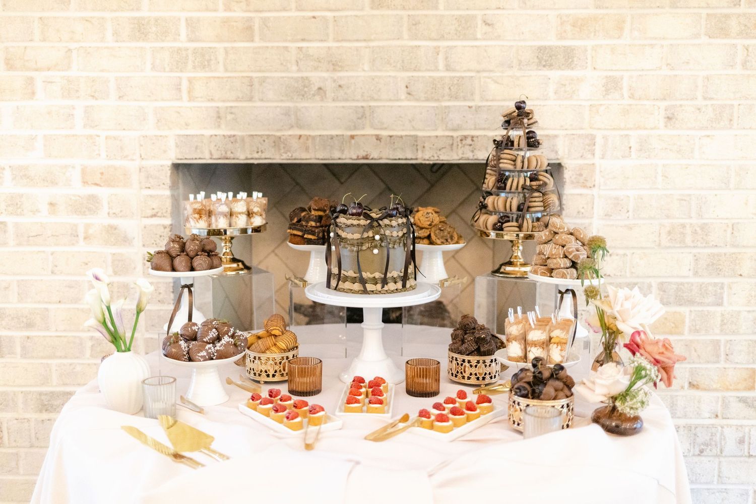An elegant dessert table display with chocolate cake, pastries, and treats arranged on a white tablecloth against a brick wall.