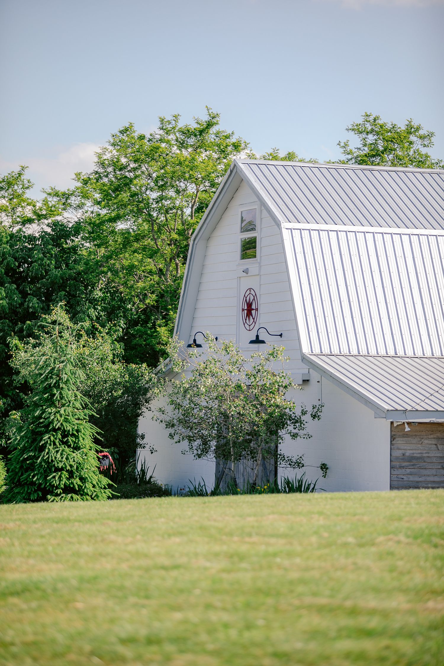 White barn with metal roof sits atop grassy hill surrounded by green trees on a sunny summer day.