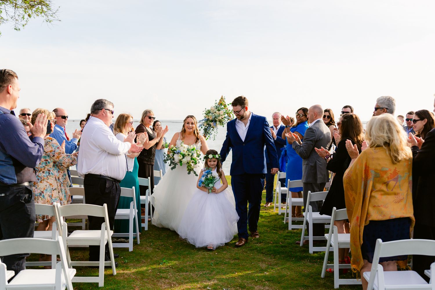 Wedding guests seated in white chairs on a lawn celebrate during an outdoor ceremony.