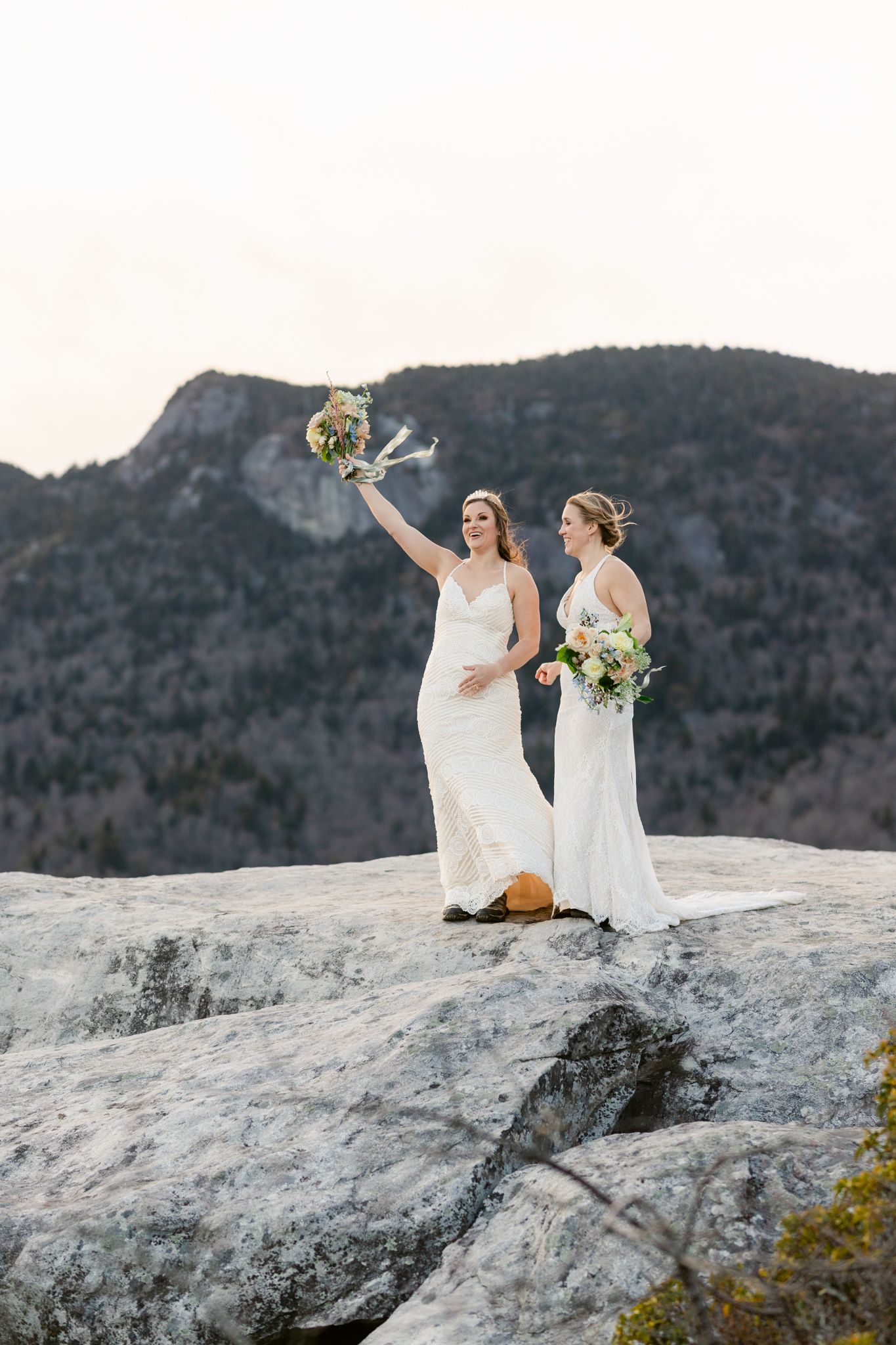 Two people in white dresses celebrate atop a scenic mountain overlook with dramatic landscape views.