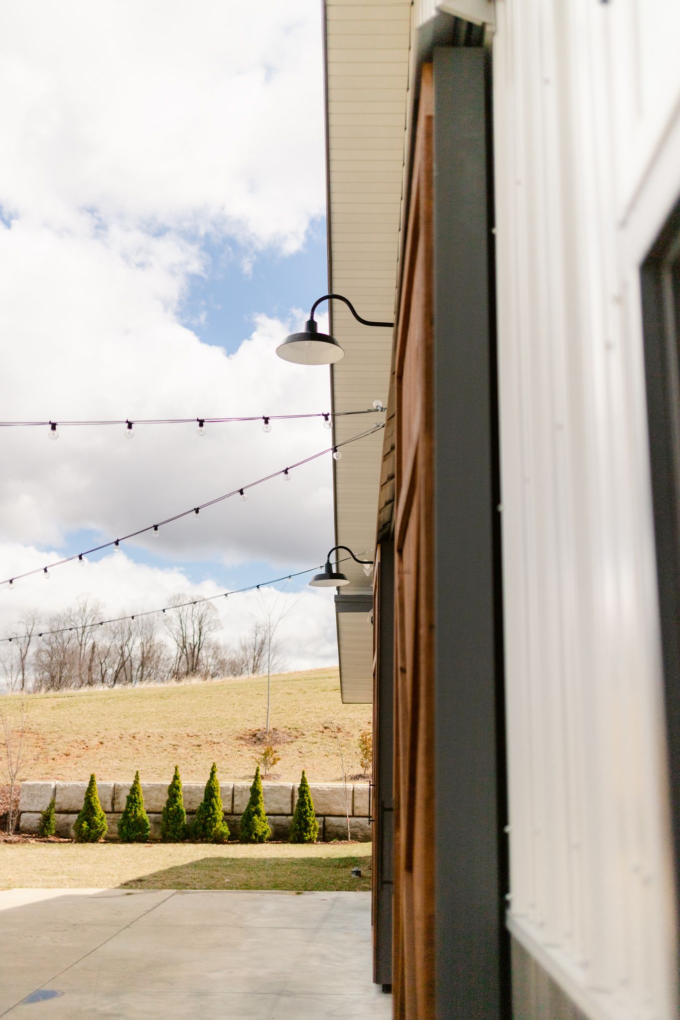 String lights hang from a modern barn exterior with white siding and black gooseneck lights against a blue sky.