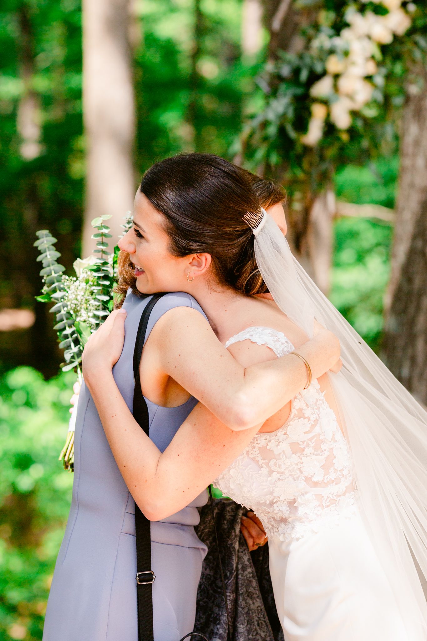 A bride in a white lace dress hugs a person wearing a gray dress in an outdoor woodland setting.