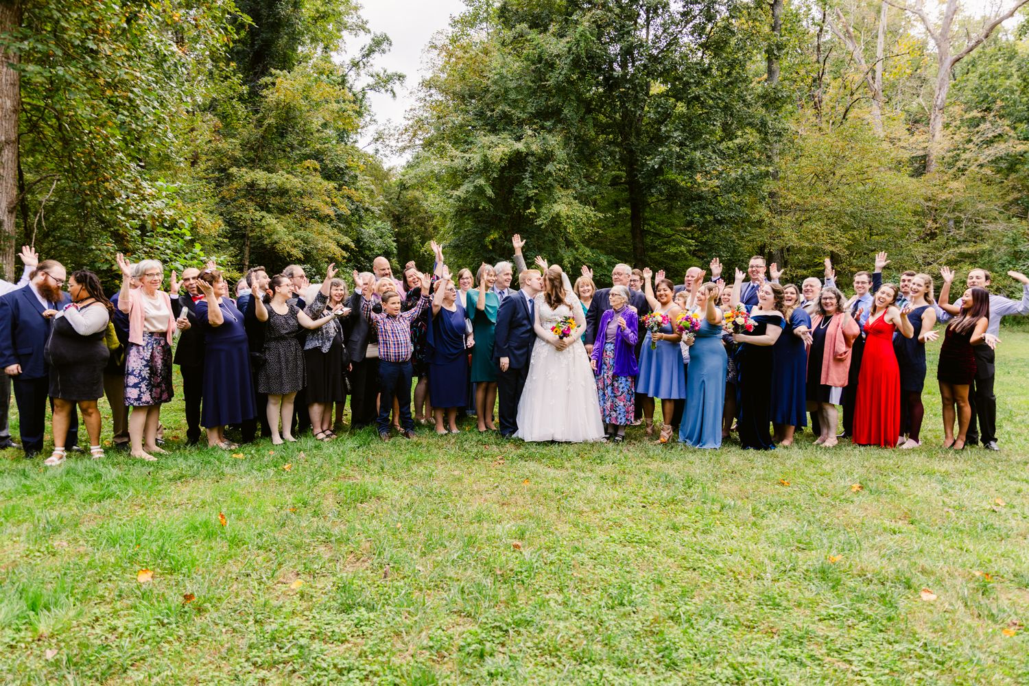 A large group of wedding guests pose together outdoors in formal attire on a grassy lawn surrounded by trees.