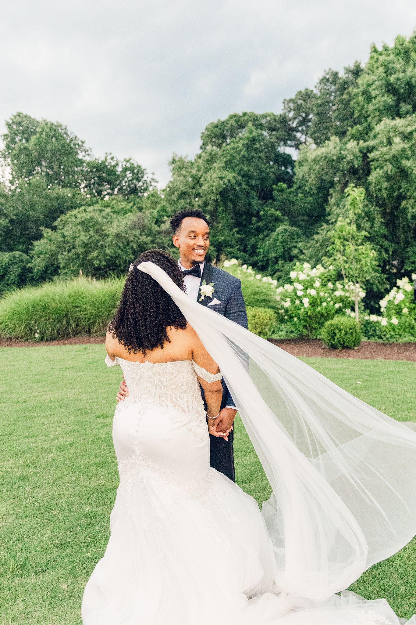 A couple shares an intimate moment on their wedding day in a lush garden setting as the bride's veil flows in the breeze.