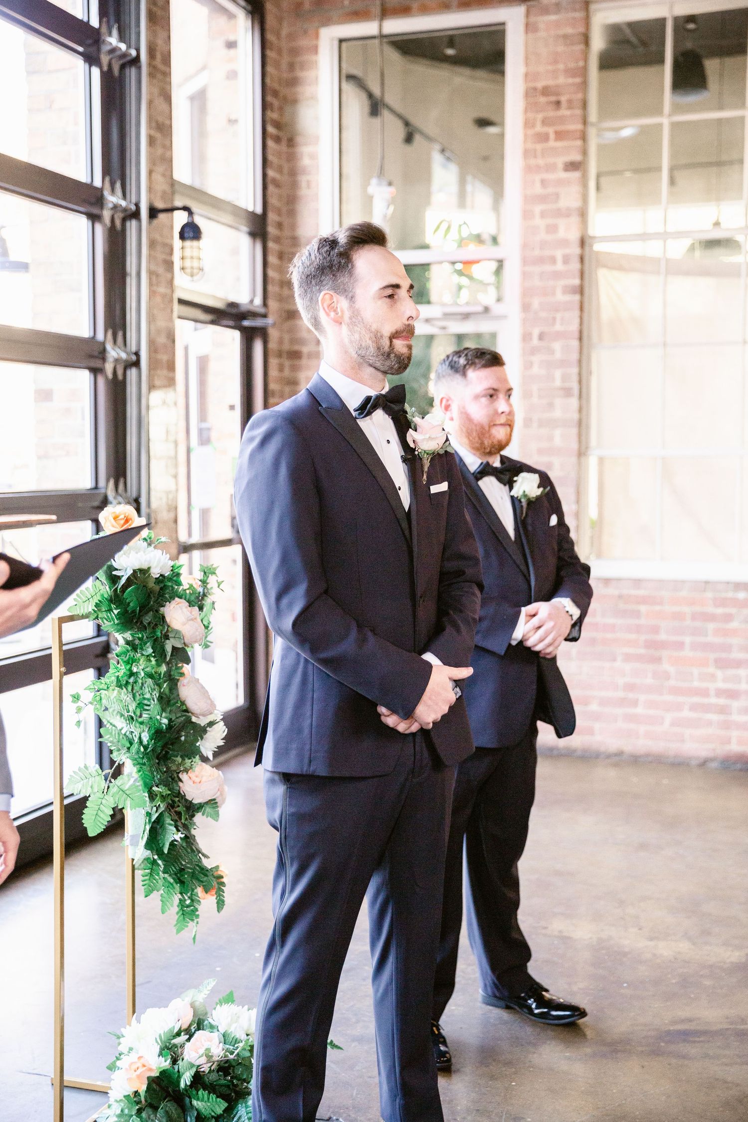 Two men in dark suits share a candid moment in a bright industrial space with brick walls and large windows.