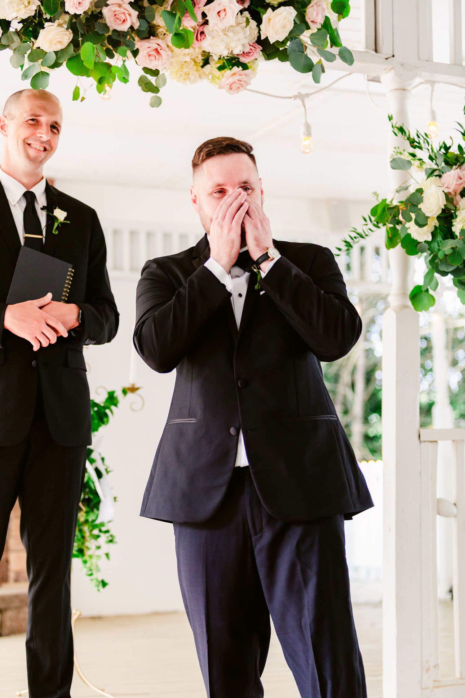 Two people in formal black suits sharing a joyful moment under floral decorations.