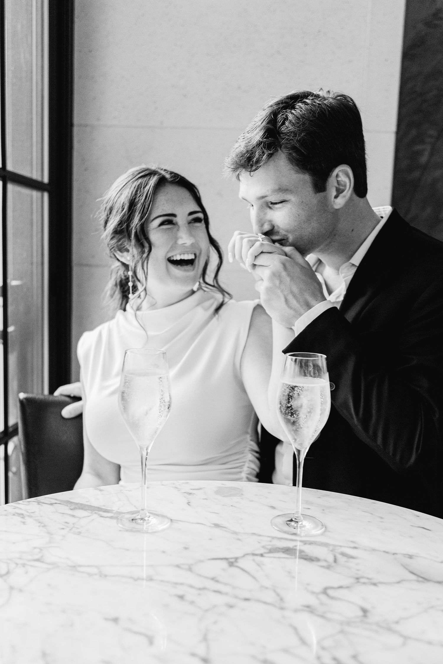 Black and white photo of a couple sharing a joyful moment at a marble table with champagne glasses.
