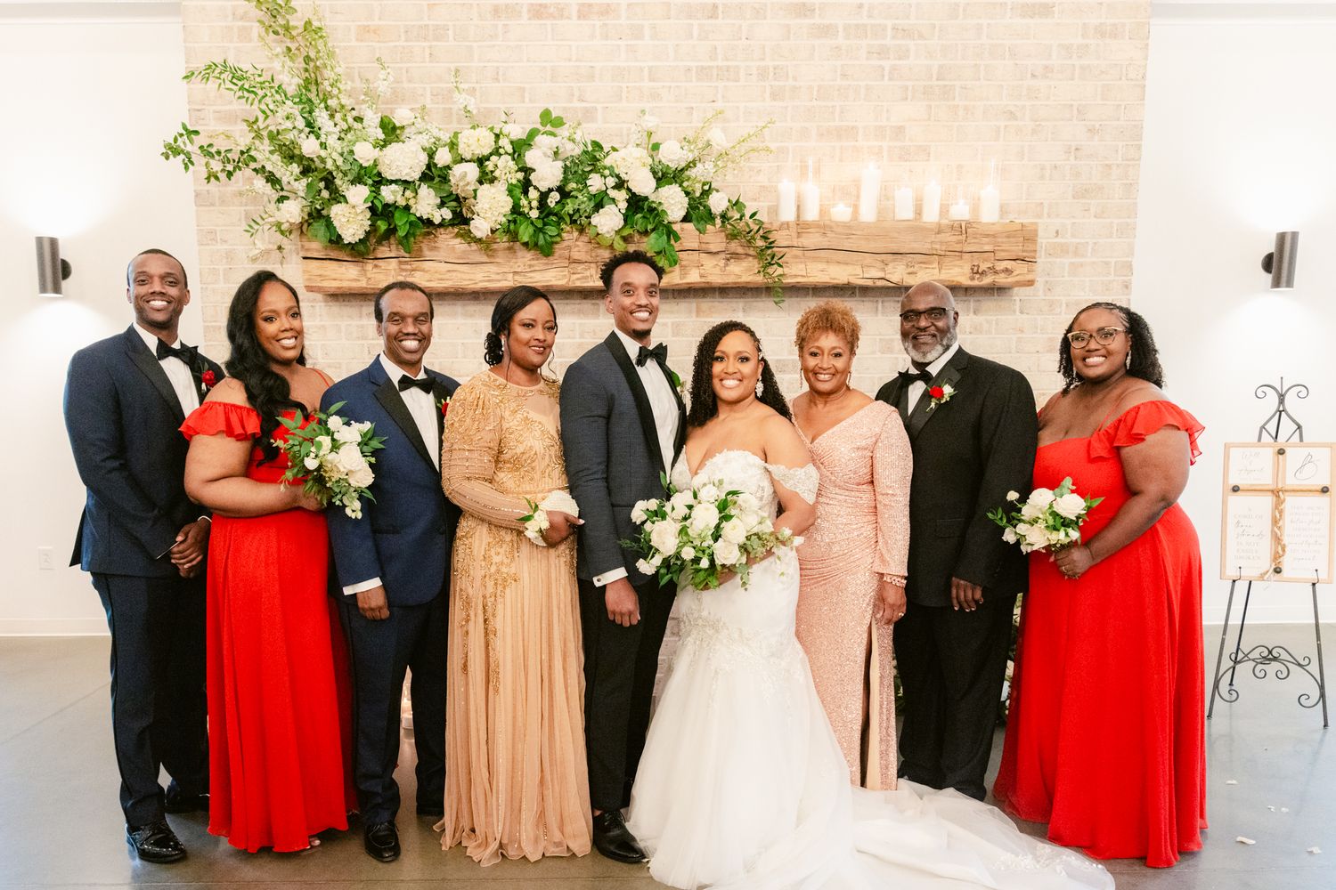 Wedding party members pose together in formal attire with red and beige dresses against a light-colored wall.