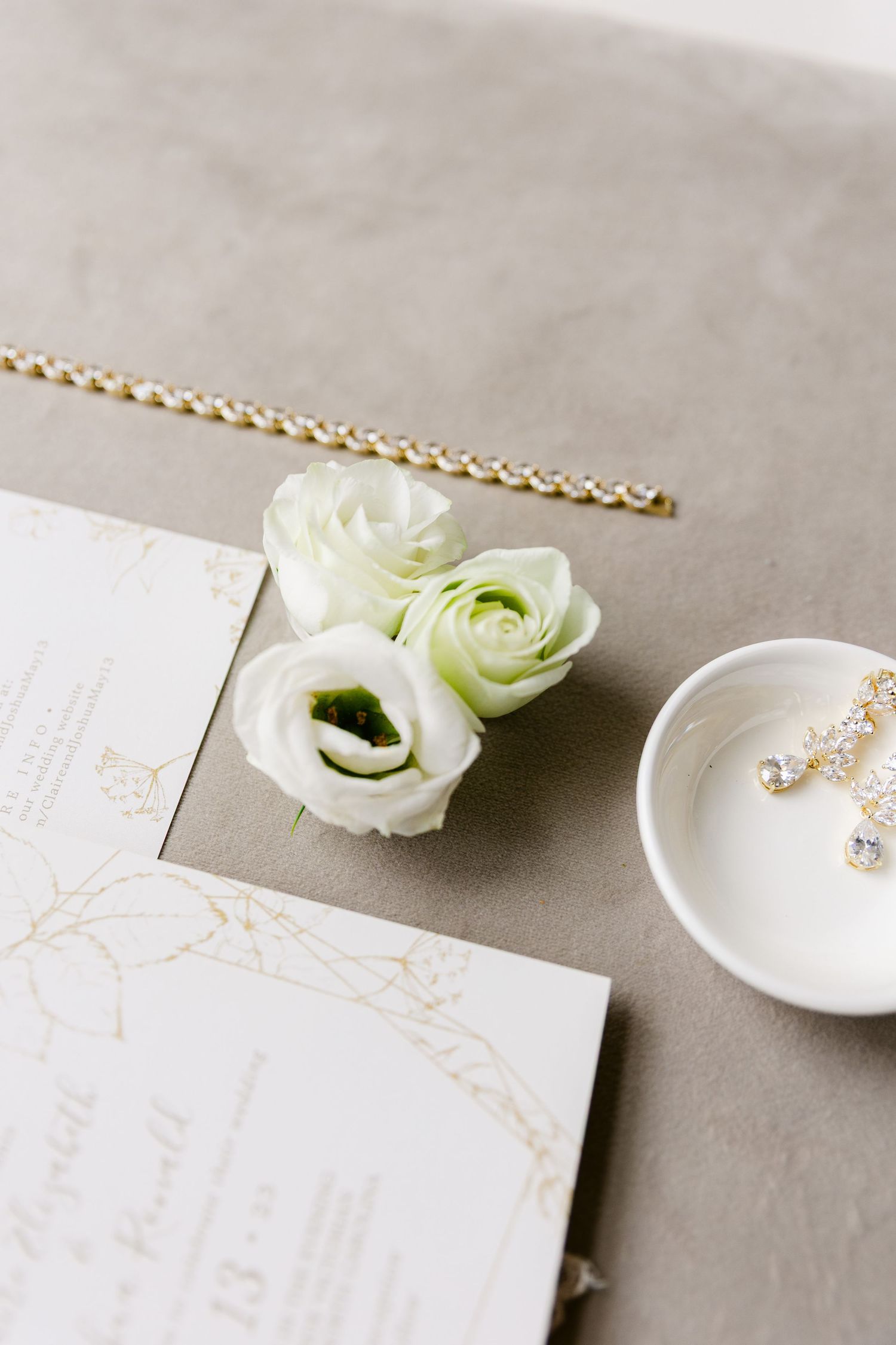 Elegant white ranunculus flowers and delicate gold jewelry arranged with minimalist white cards on a neutral gray surface.