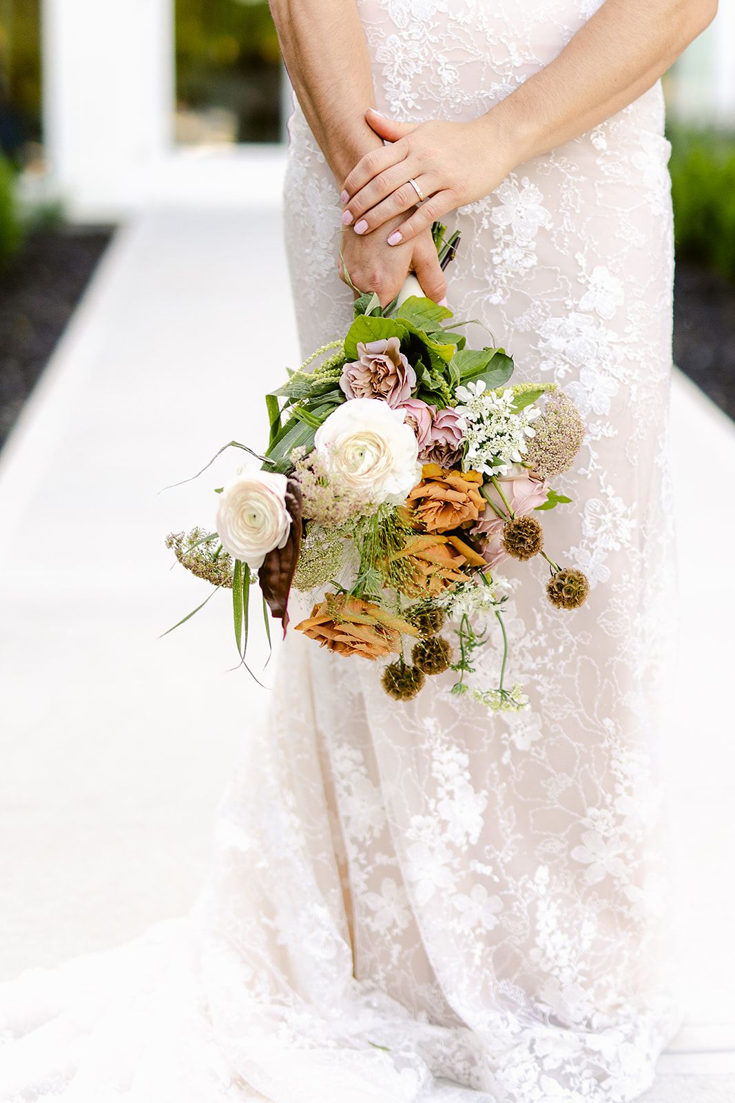 Close-up detail of a delicate white lace wedding dress with a rustic floral bouquet featuring roses and greenery.
