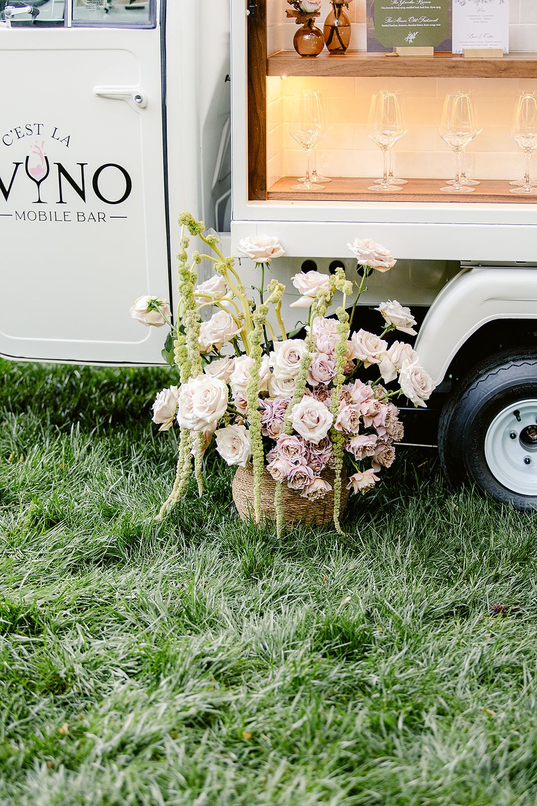 Beautiful floral arrangement with pale pink roses and white flowers sits beside a mobile bar truck on green grass.