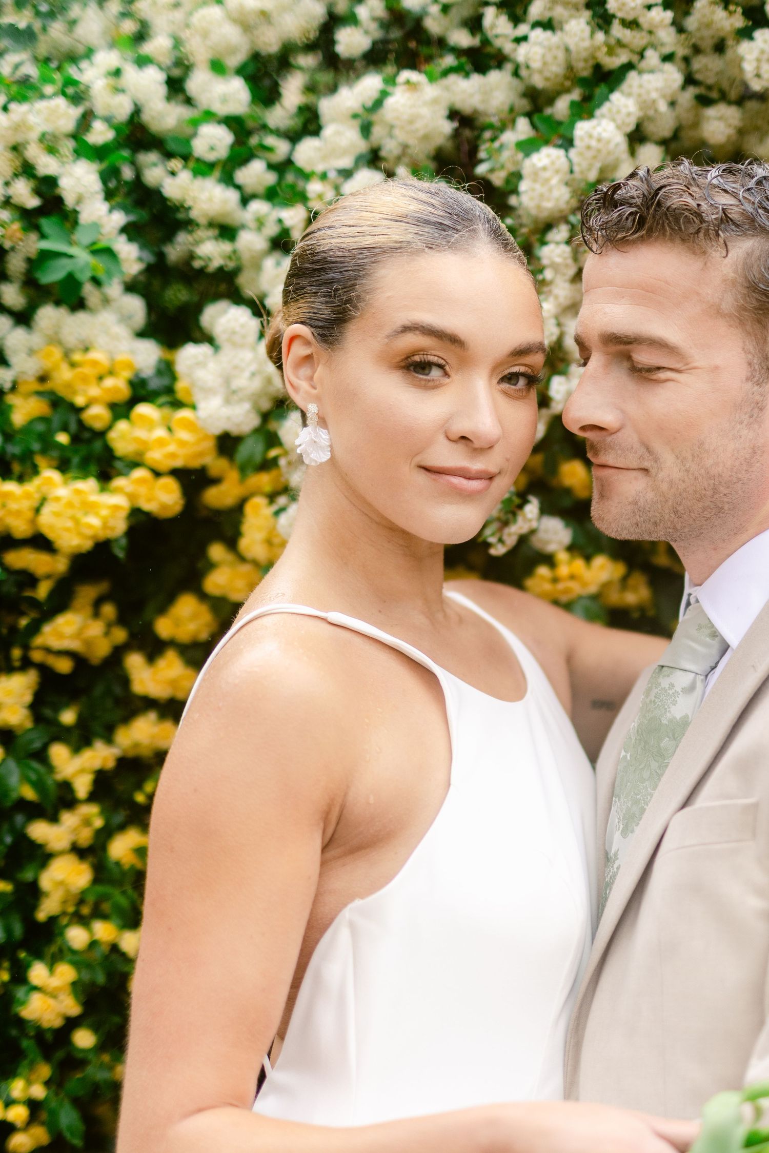 A romantic couple shares an intimate moment in front of a backdrop of white and yellow flowers in a garden setting.