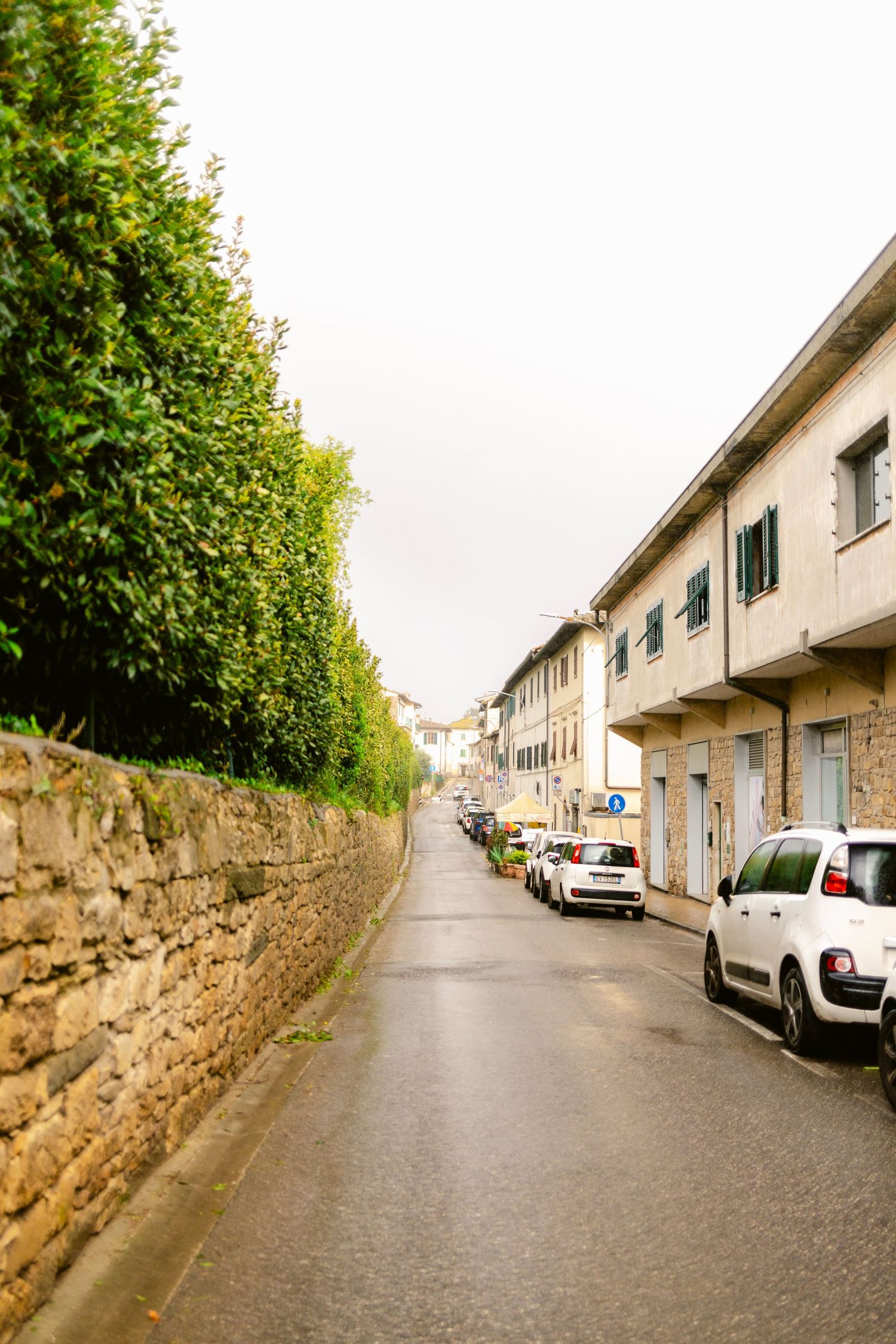 Narrow residential street lined with parked cars and a stone wall leading to row houses in the distance.