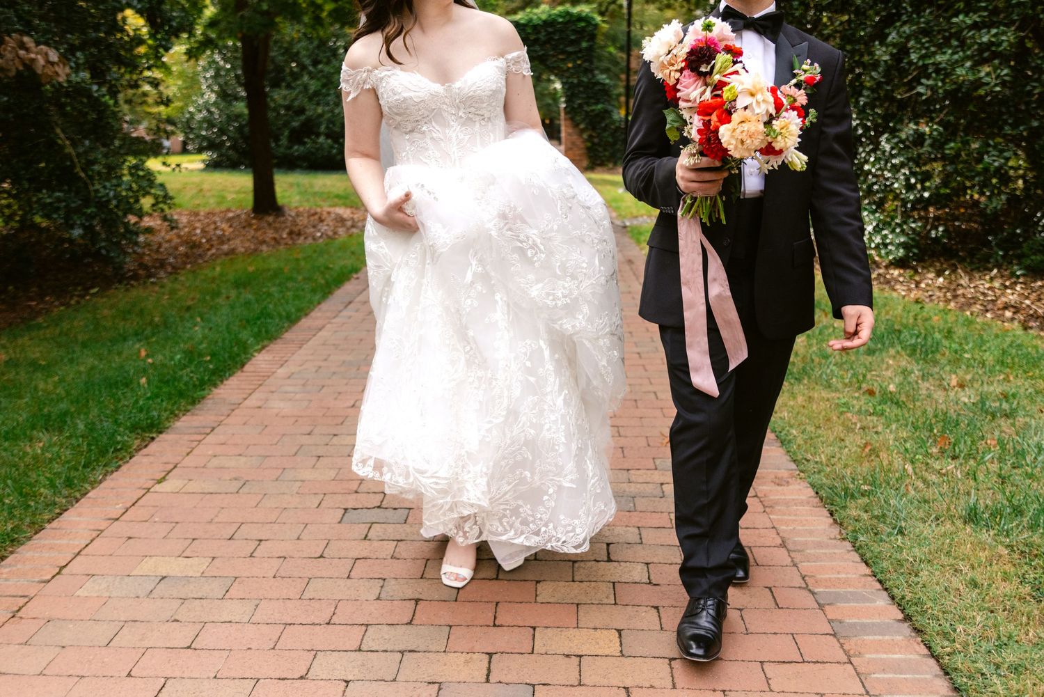 A couple walks down a brick path surrounded by lush greenery, with the bride's white dress flowing behind them.