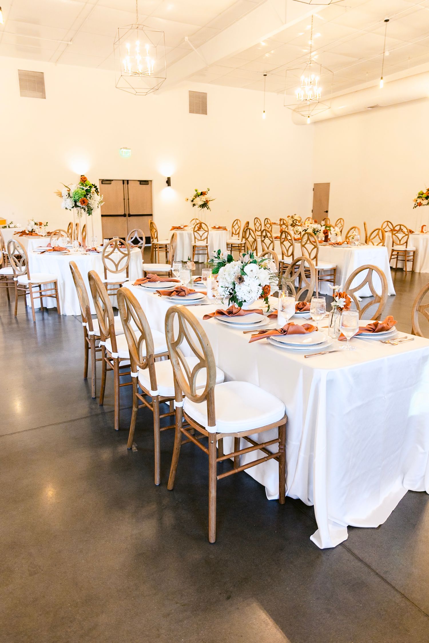 Wedding reception venue setup with elegant white tablecloths and wooden cross-back chairs in a modern industrial space.