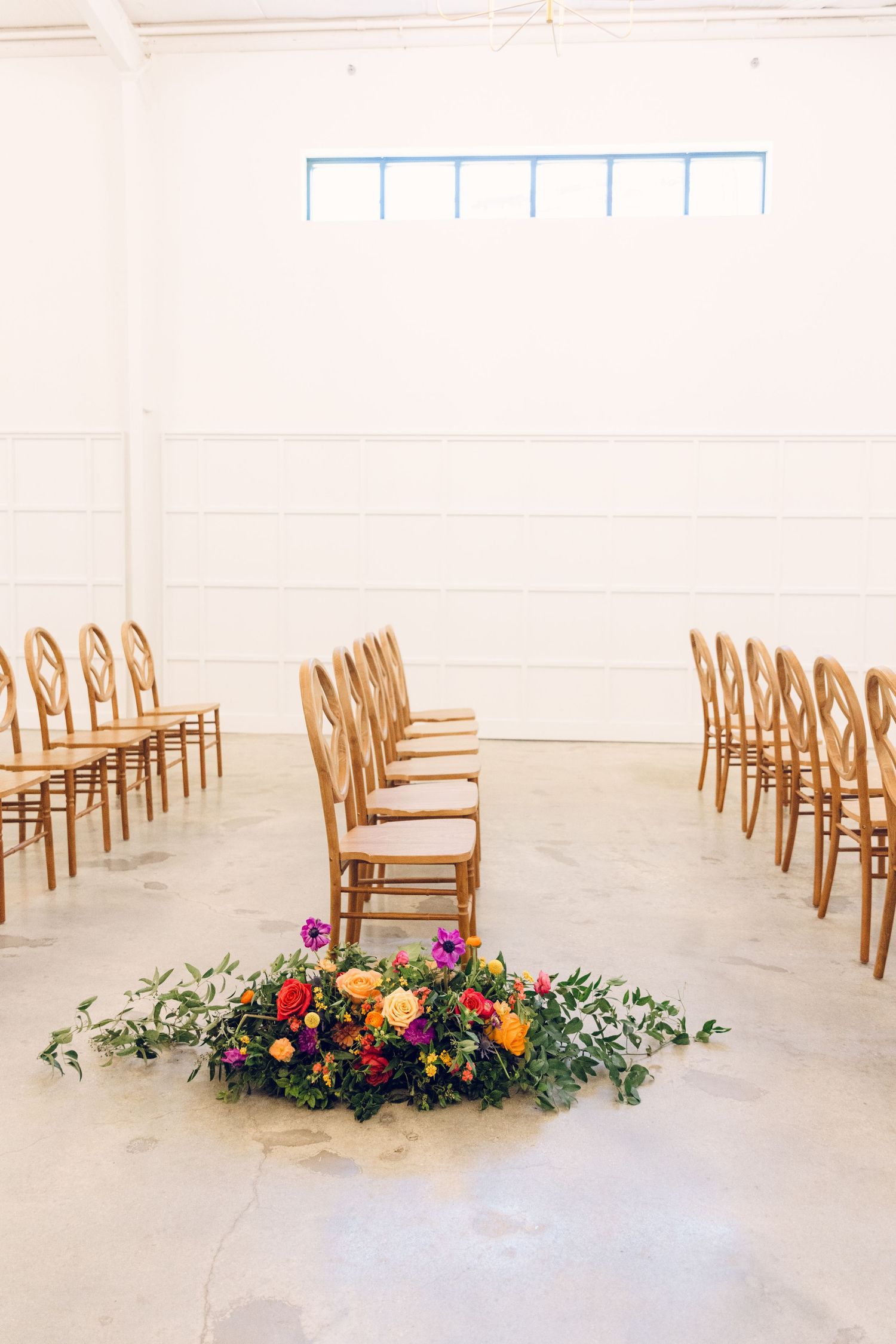 Wooden chairs arranged in rows with colorful floral arrangements lining the aisle in a minimalist white ceremony space.