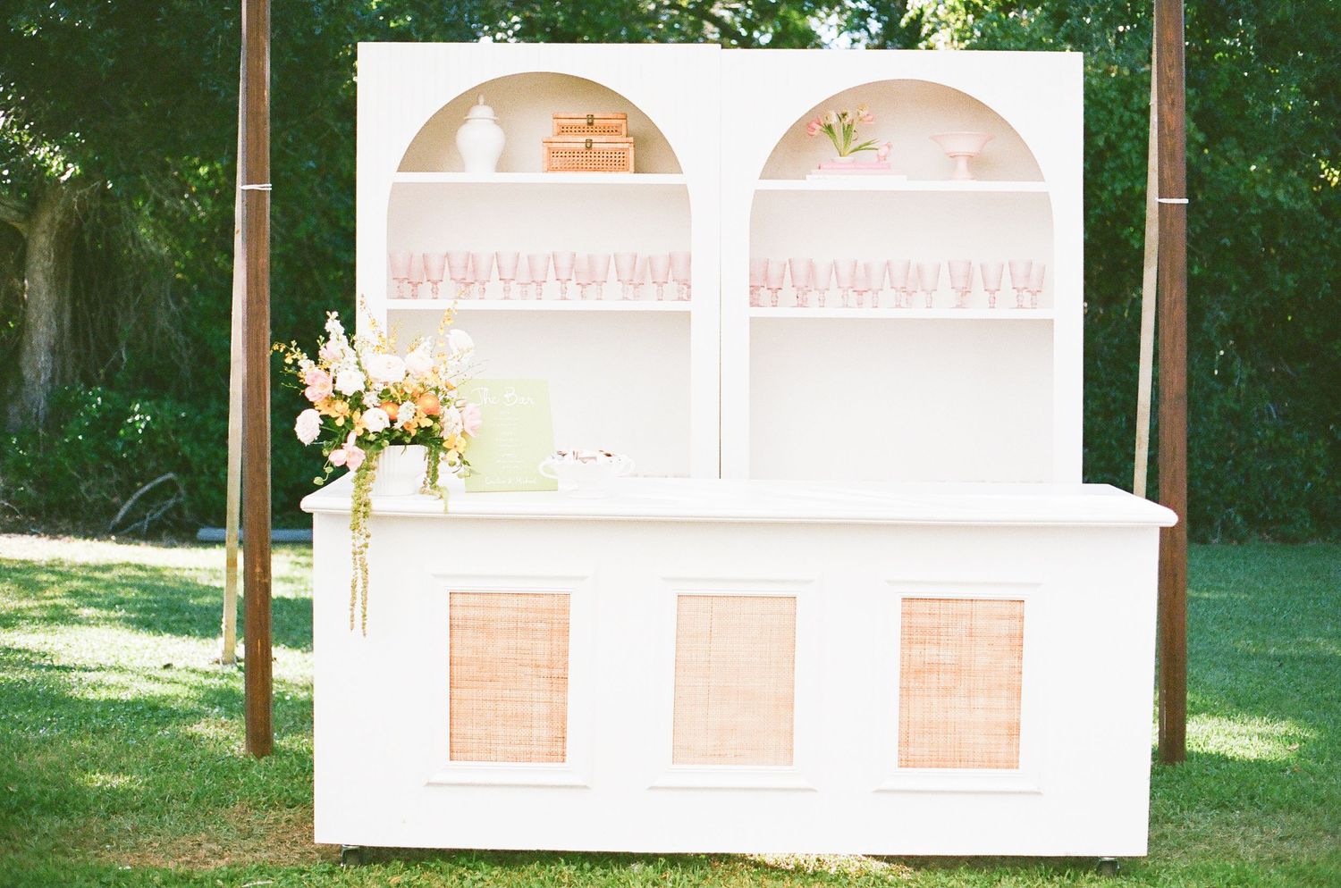 White vintage-style bar cabinet with arched shelving displays glassware at outdoor wedding.