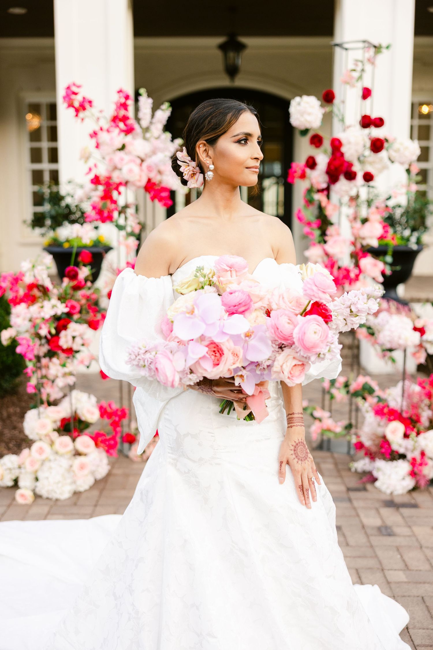 A bride in a white gown poses elegantly among pink floral arrangements and white columns.
