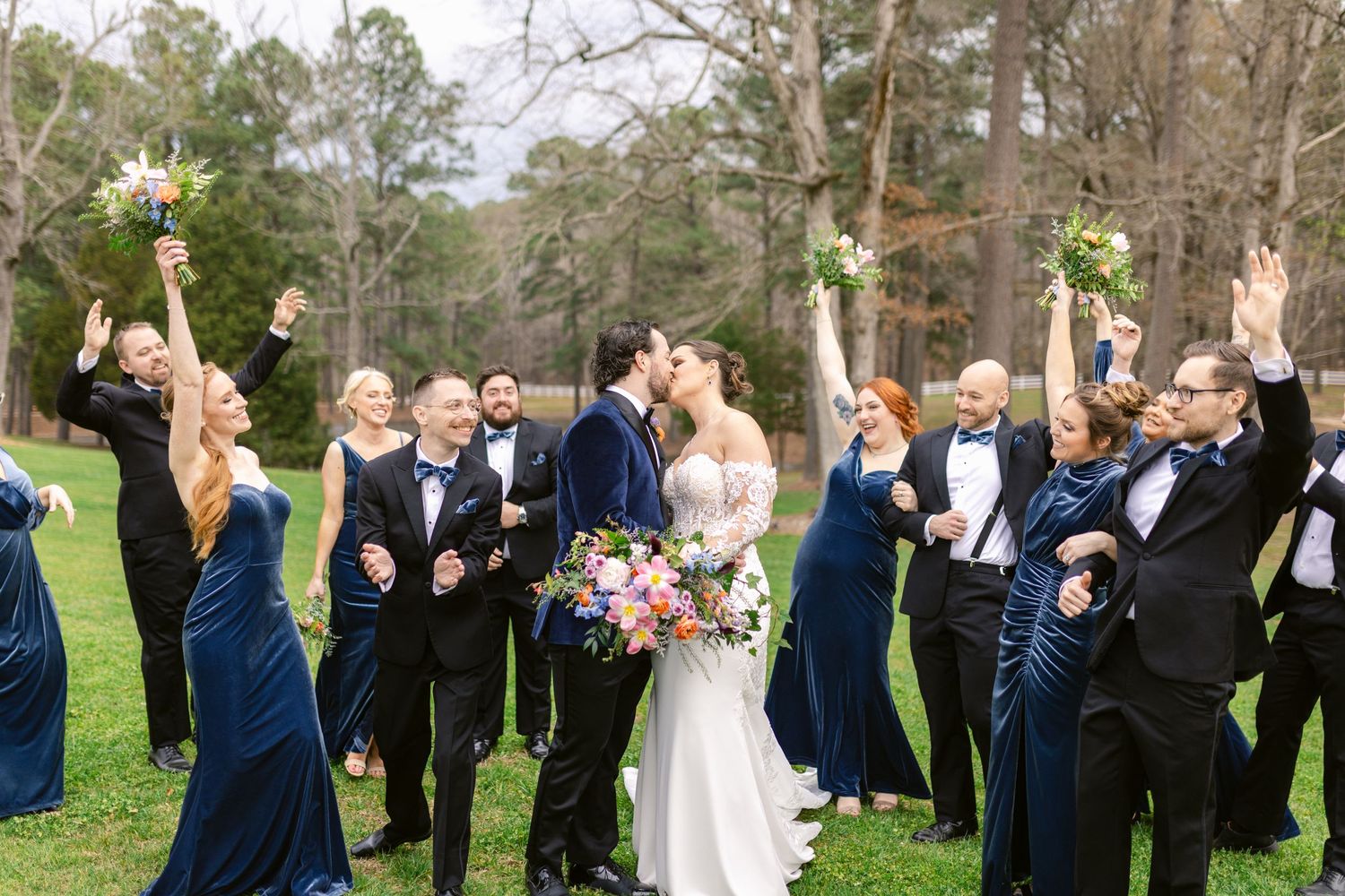 Wedding party in navy blue and white attire celebrating joyfully on a green lawn with trees in the background.