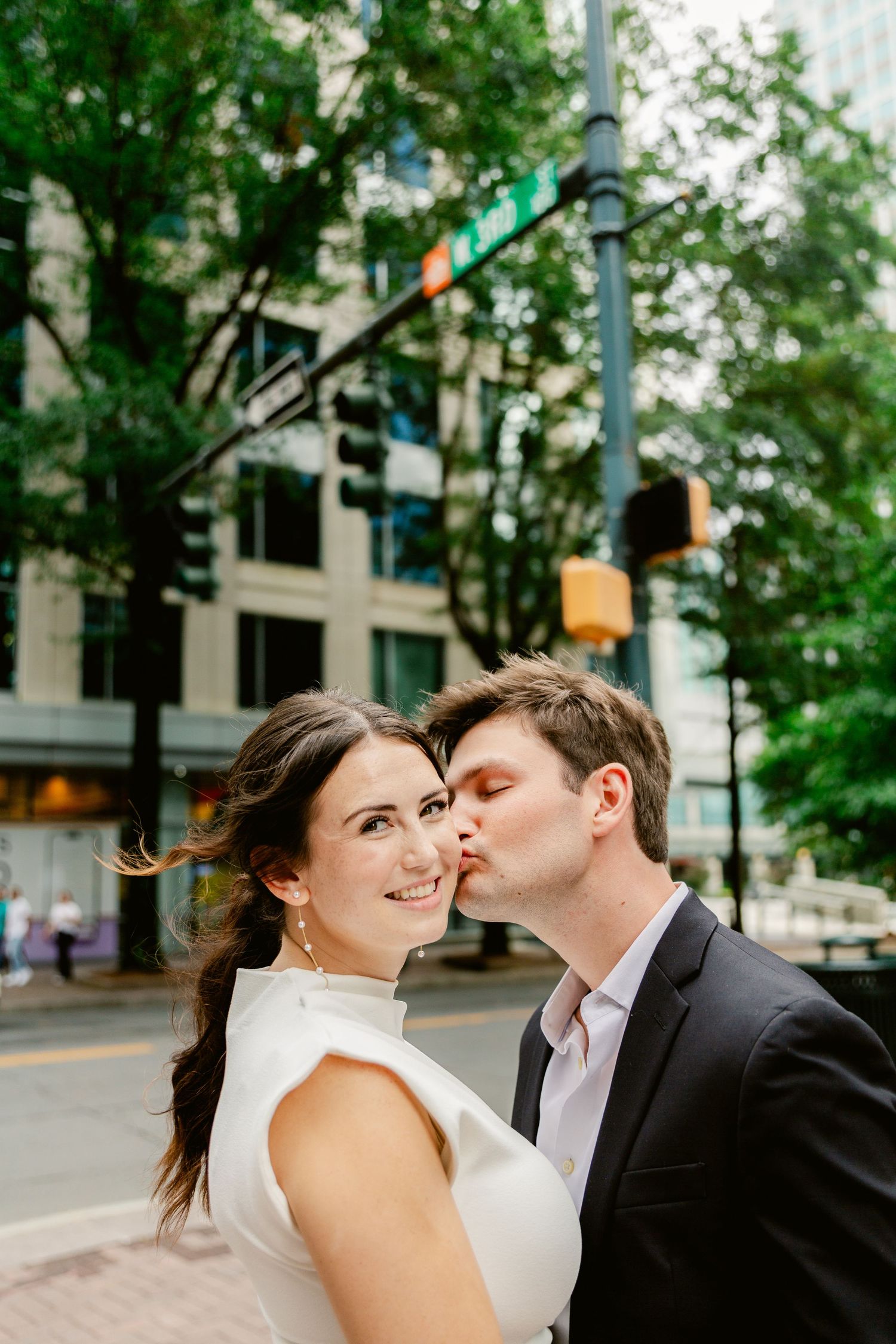 Couple shares romantic moment on city street corner with traffic light and trees in urban setting.