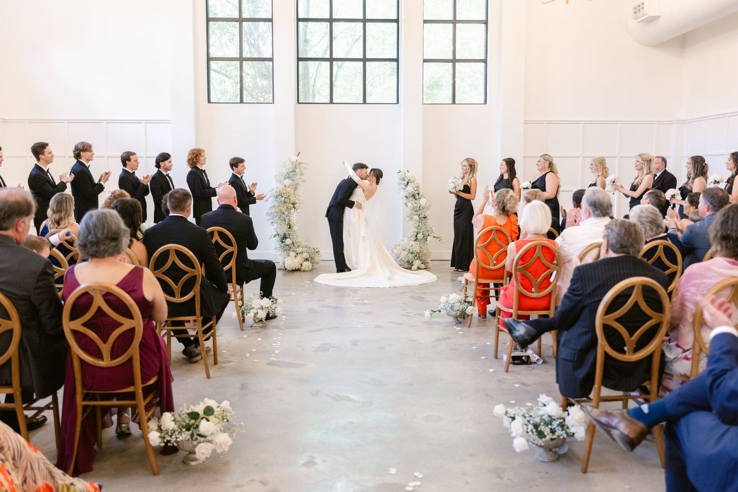 Wedding ceremony in a bright white room with gold chairs and white floral arrangements framing the aisle.