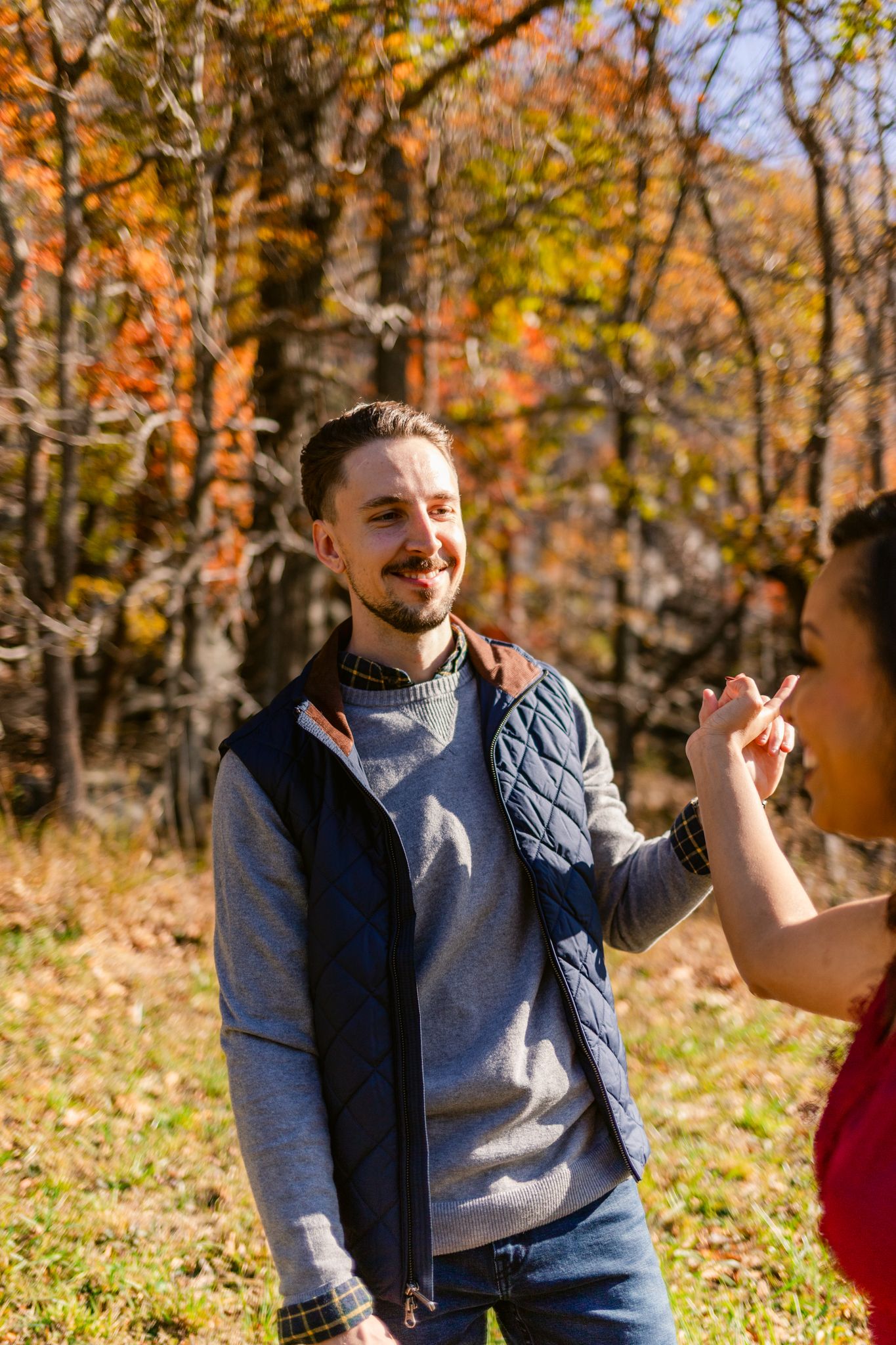 Person wearing a quilted vest and grey sweater smiles while standing in a colorful autumn forest setting.
