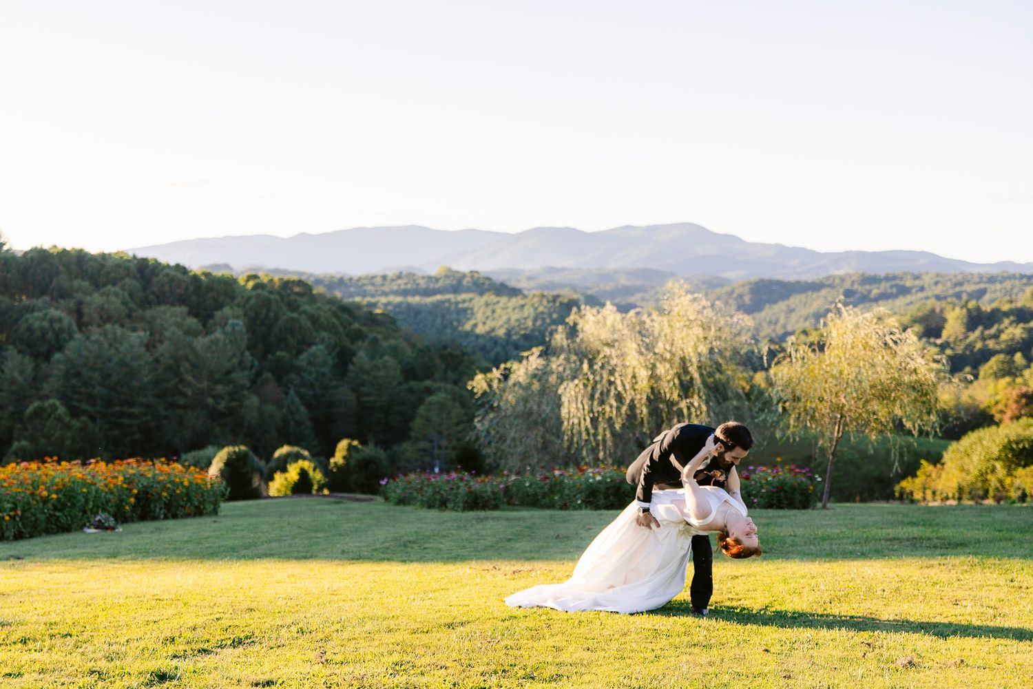 A couple shares a romantic moment on a hilltop meadow overlooking mountains and forests at golden hour.