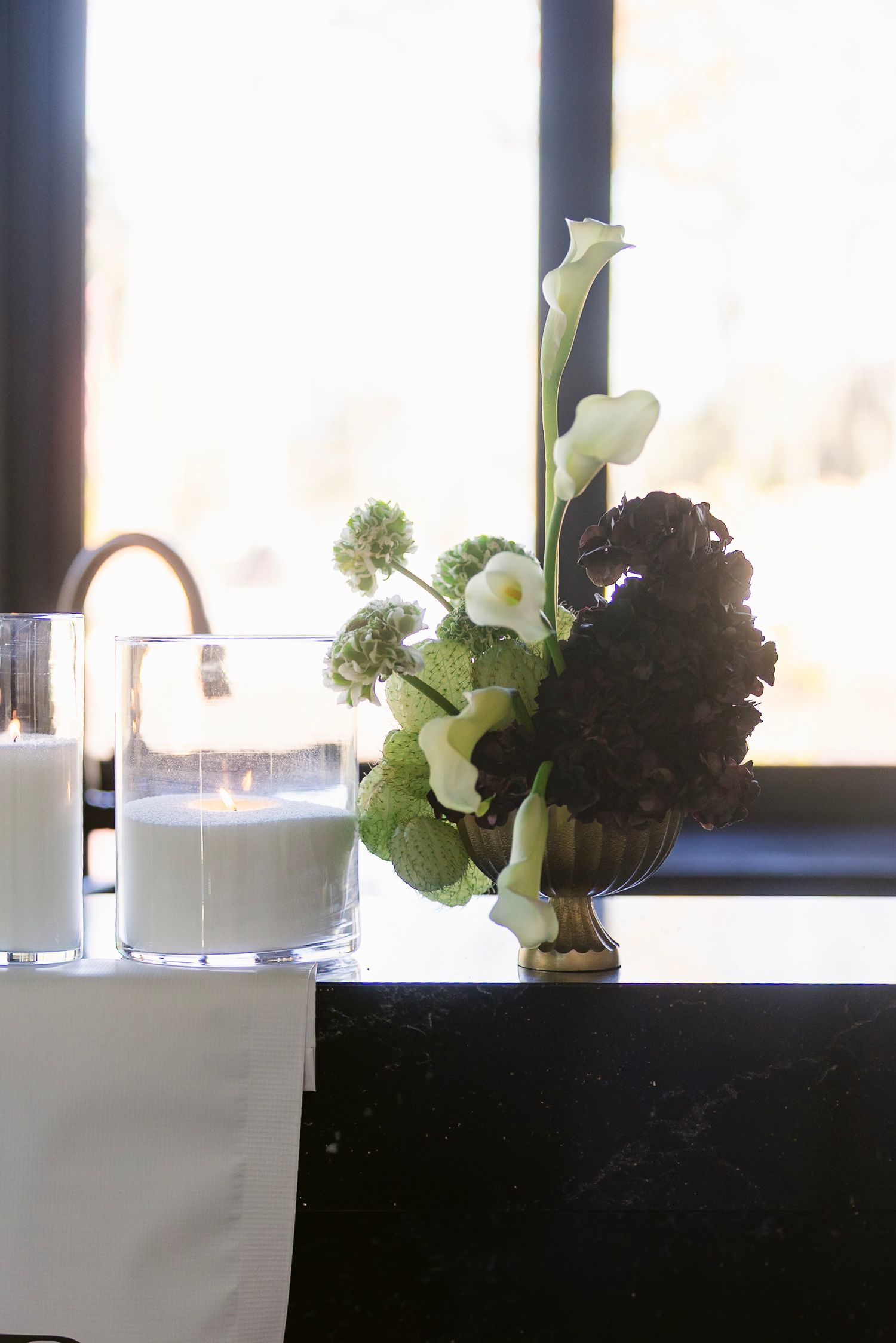 Elegant white floral arrangements in cylindrical vases line a modern black counter with window lighting.