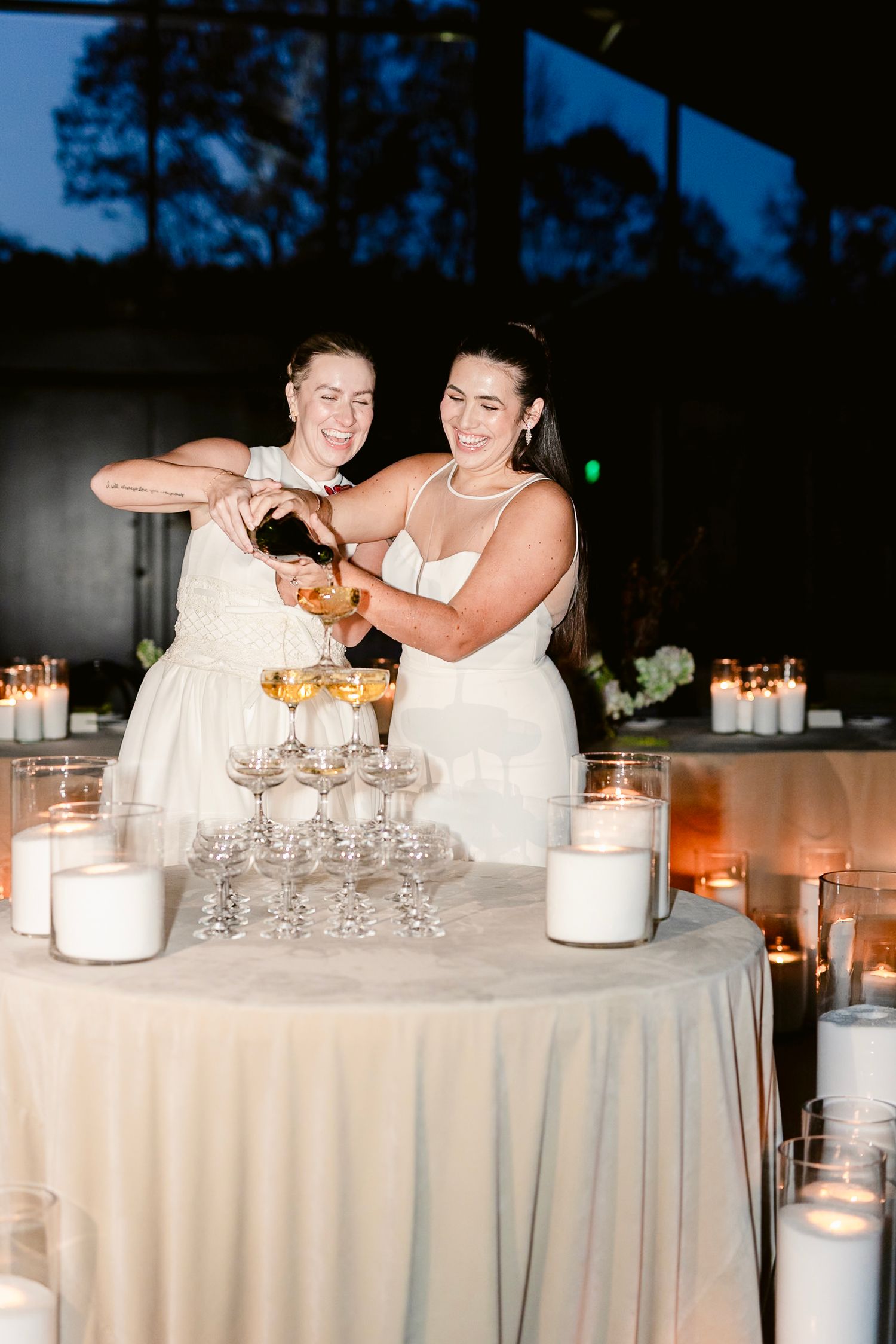 Two people in white dresses share a laugh while seated at an elegantly set table with wine glasses and candles.