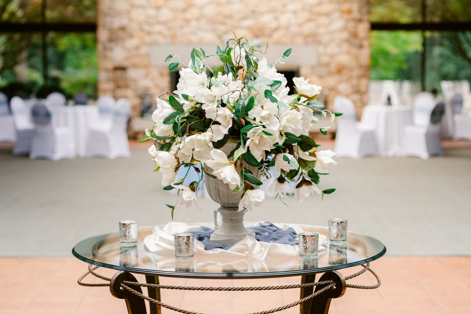 Elegant white floral centerpiece arrangement on mirrored table with reception tables and chairs in background.