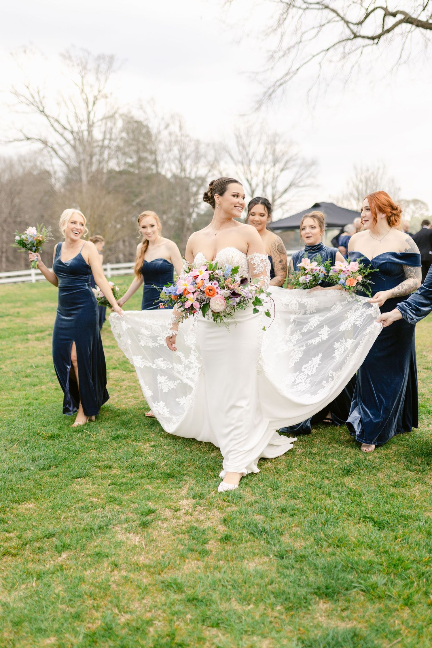 Bridesmaids in navy blue dresses help adjust a flowing white wedding gown on a grassy lawn during an outdoor celebration.