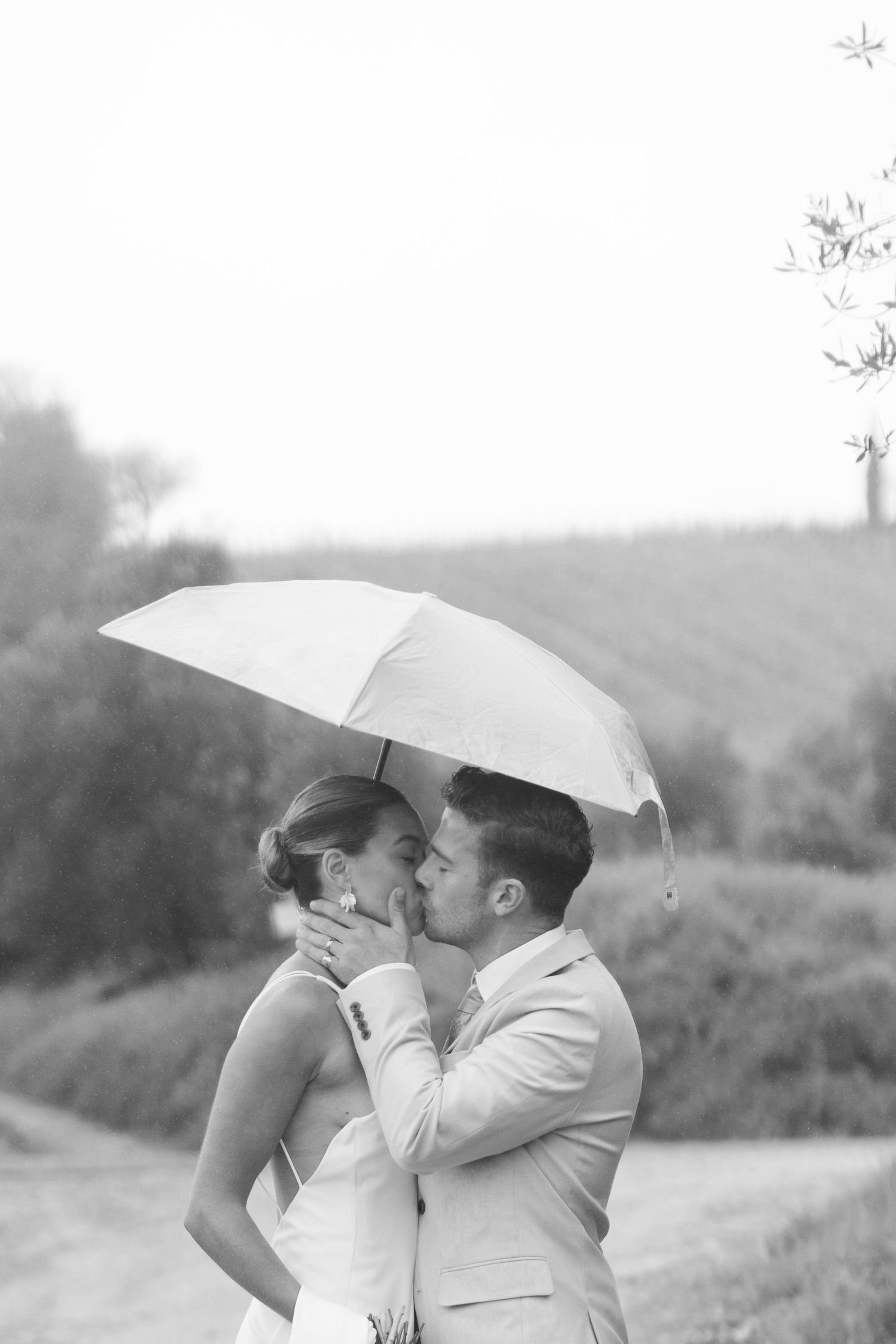 A romantic couple shares a kiss under a white umbrella in a dreamy black and white outdoor setting.