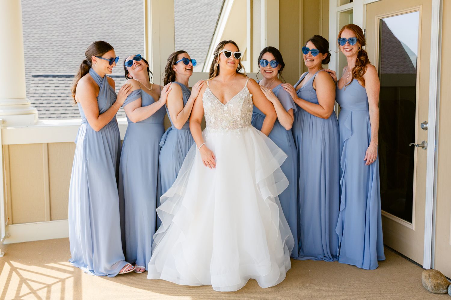 A wedding party in light blue bridesmaid dresses poses with the bride in a white gown at a doorway.