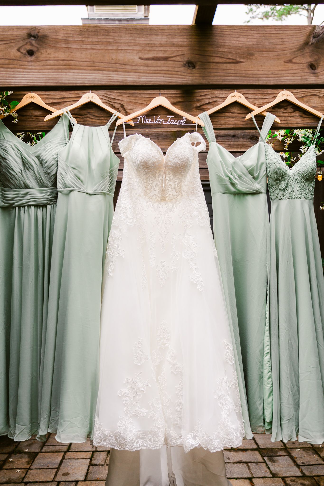 Wedding dresses in white and sage green bridesmaid gowns hanging on rustic wooden wall for display.