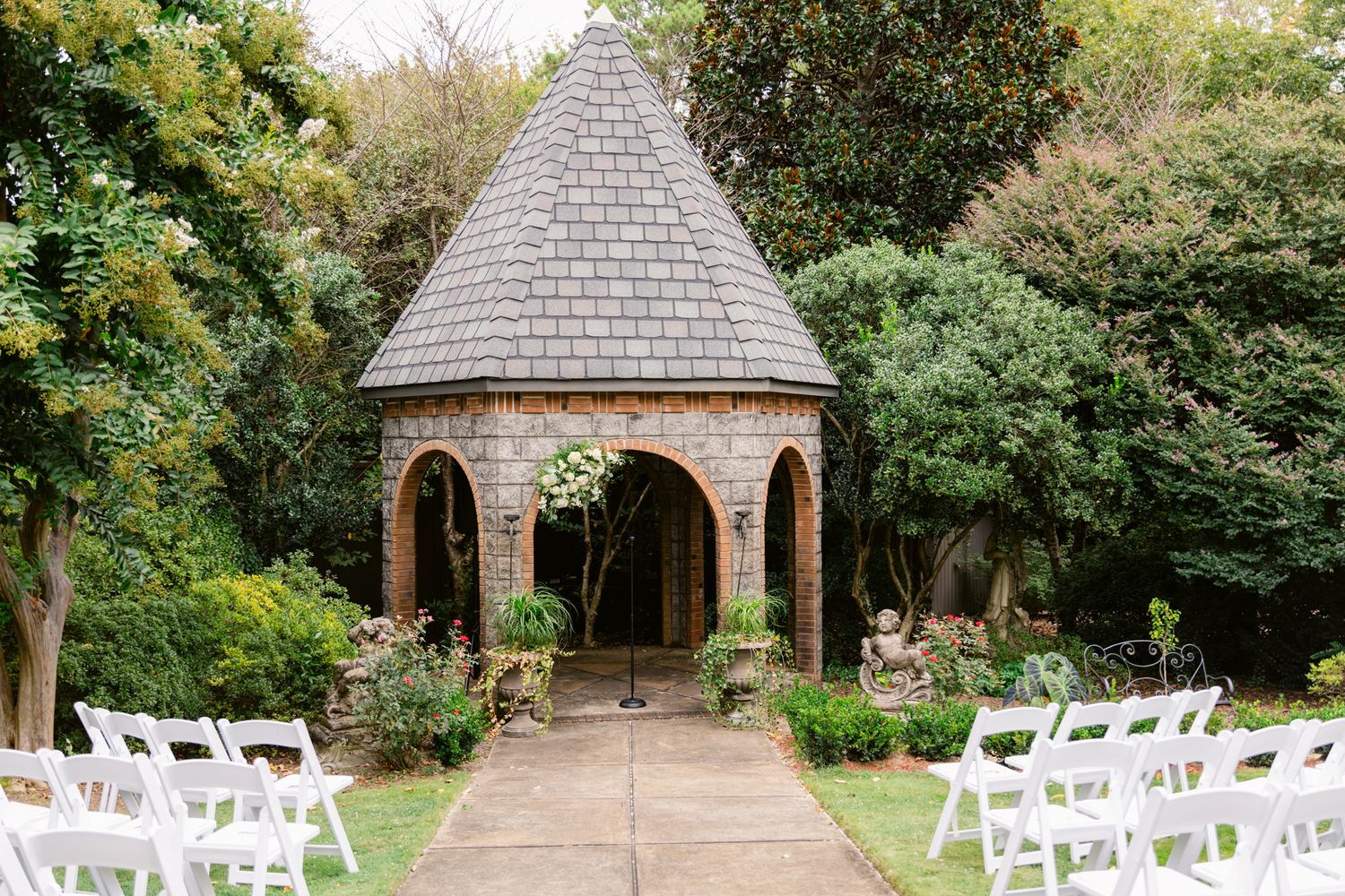 A picturesque garden wedding venue with white chairs set up in front of a stone gazebo surrounded by lush greenery.