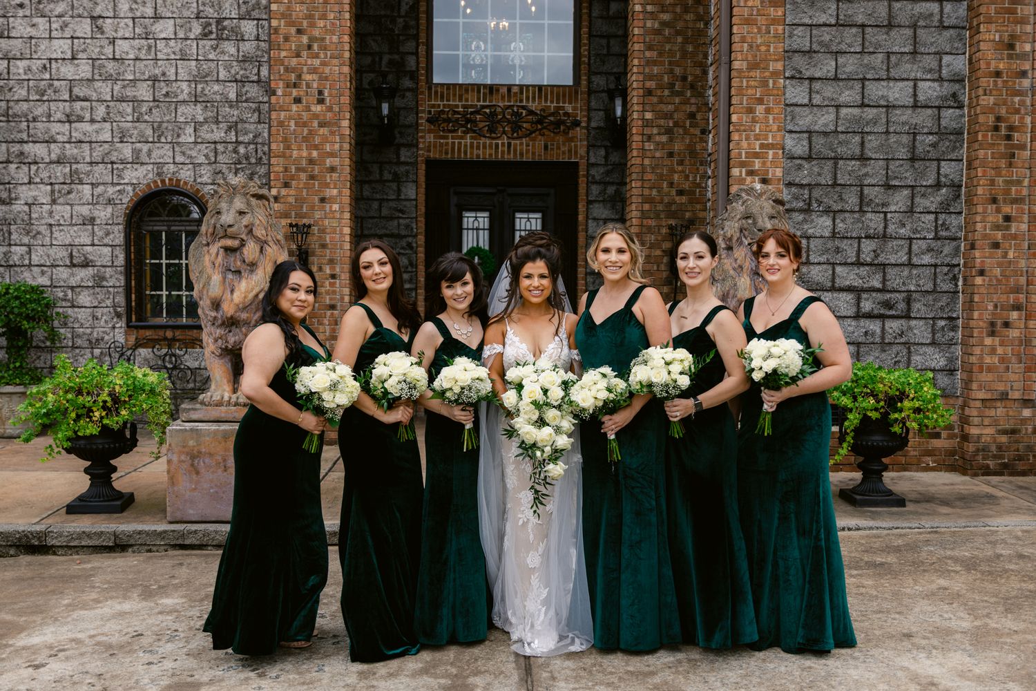 Wedding party poses against brick wall backdrop in coordinated dark green and black formal attire.