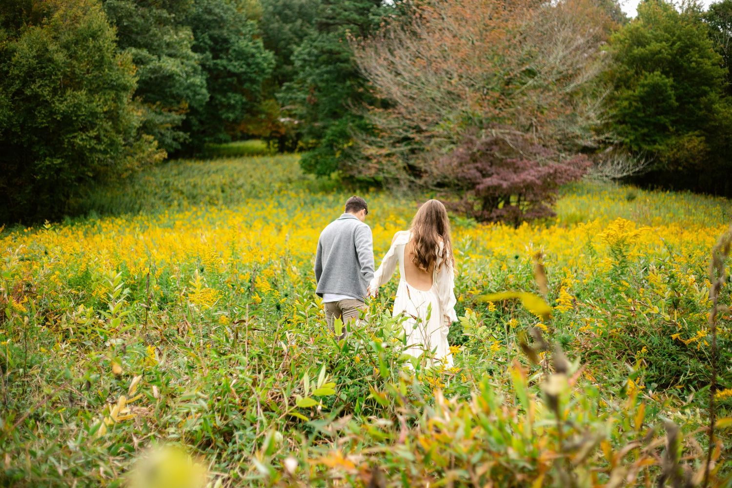 A couple walking through a vibrant field of yellow wildflowers surrounded by autumn trees.