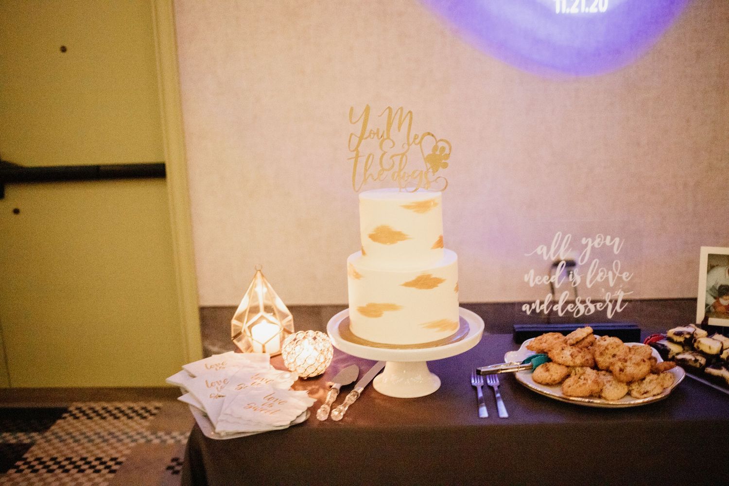 Wedding cake table with three-tier white cake and 'Mr & Mrs' topper under purple lighting.