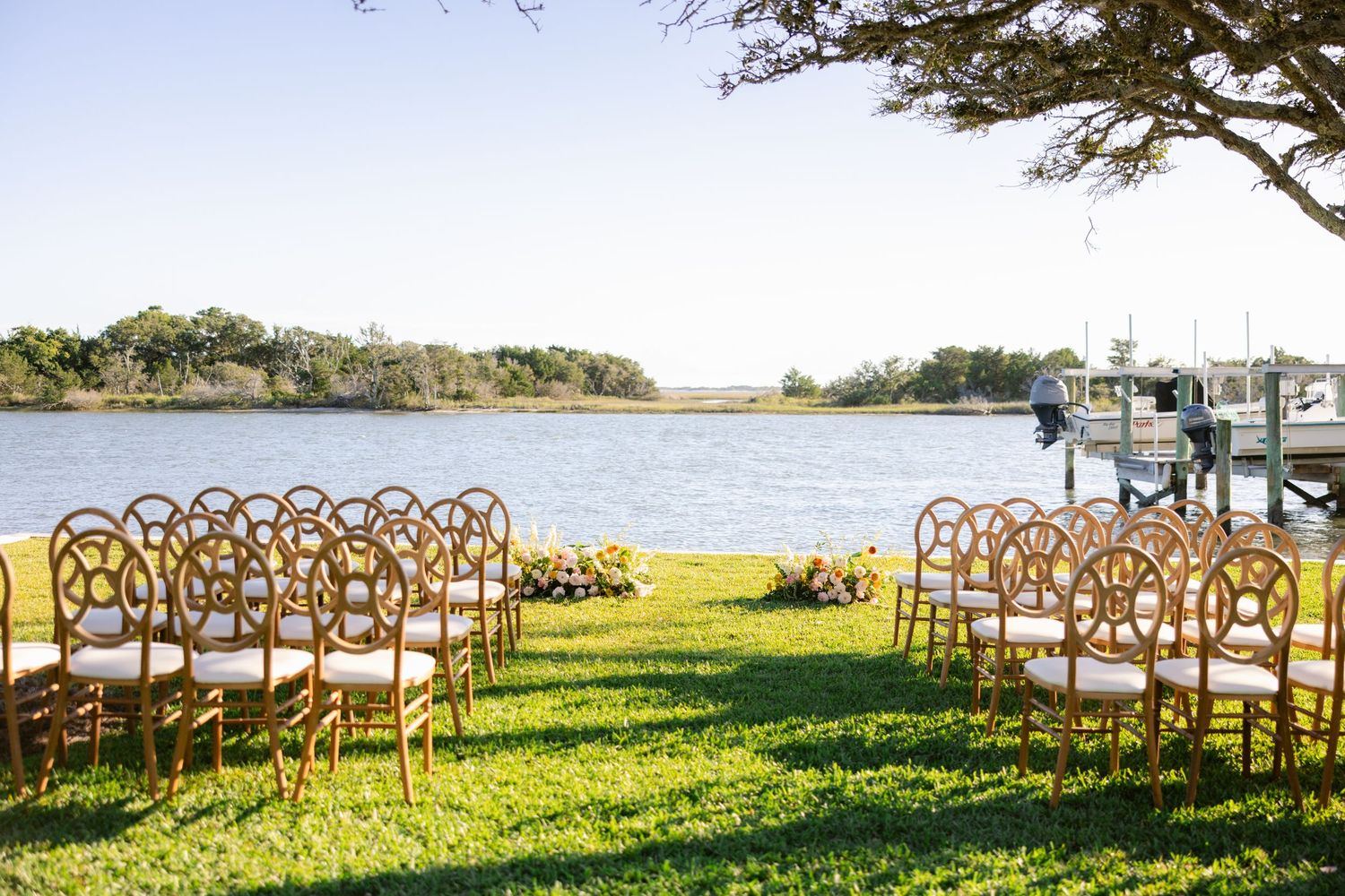 Wooden cross-back chairs arranged in rows on a grassy lawn overlooking a waterfront for an outdoor wedding ceremony.