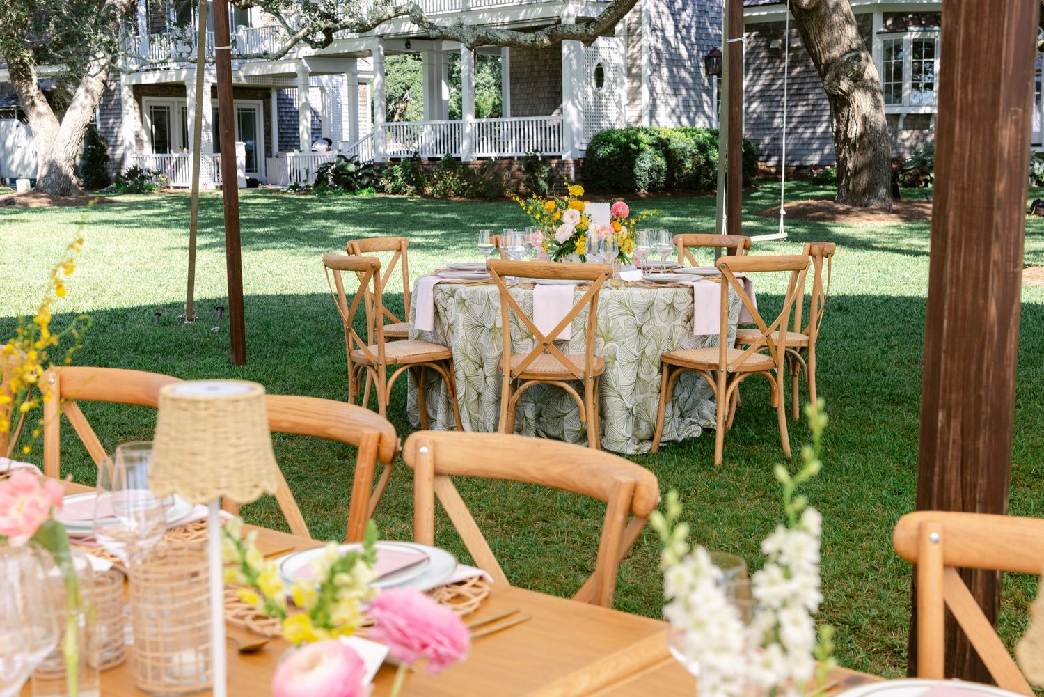 A garden wedding reception setup with wooden cross-back chairs and floral table settings under trees.