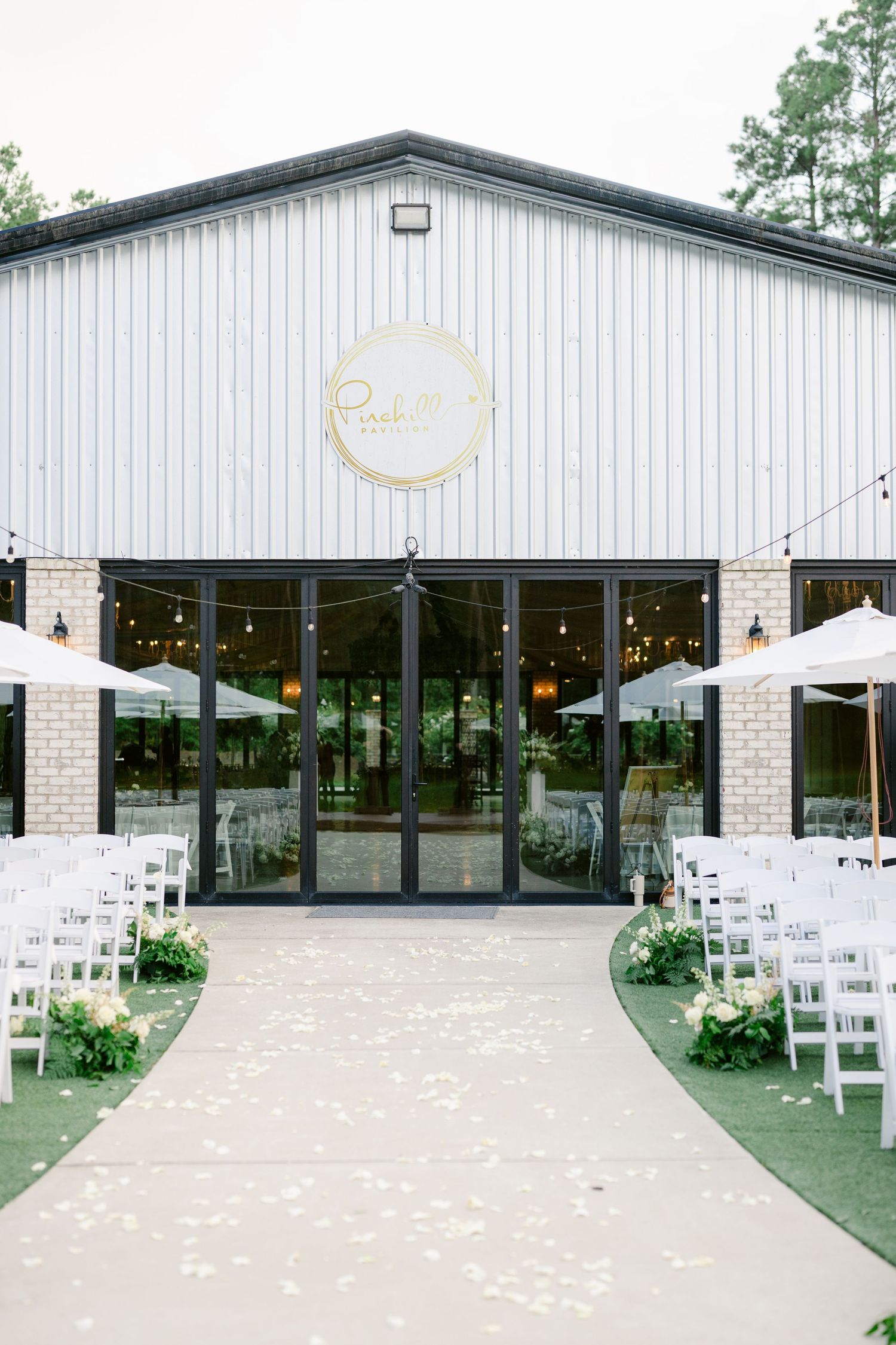 Modern white wedding venue with rustic metal buildings and white chairs set up for an outdoor ceremony on turf.