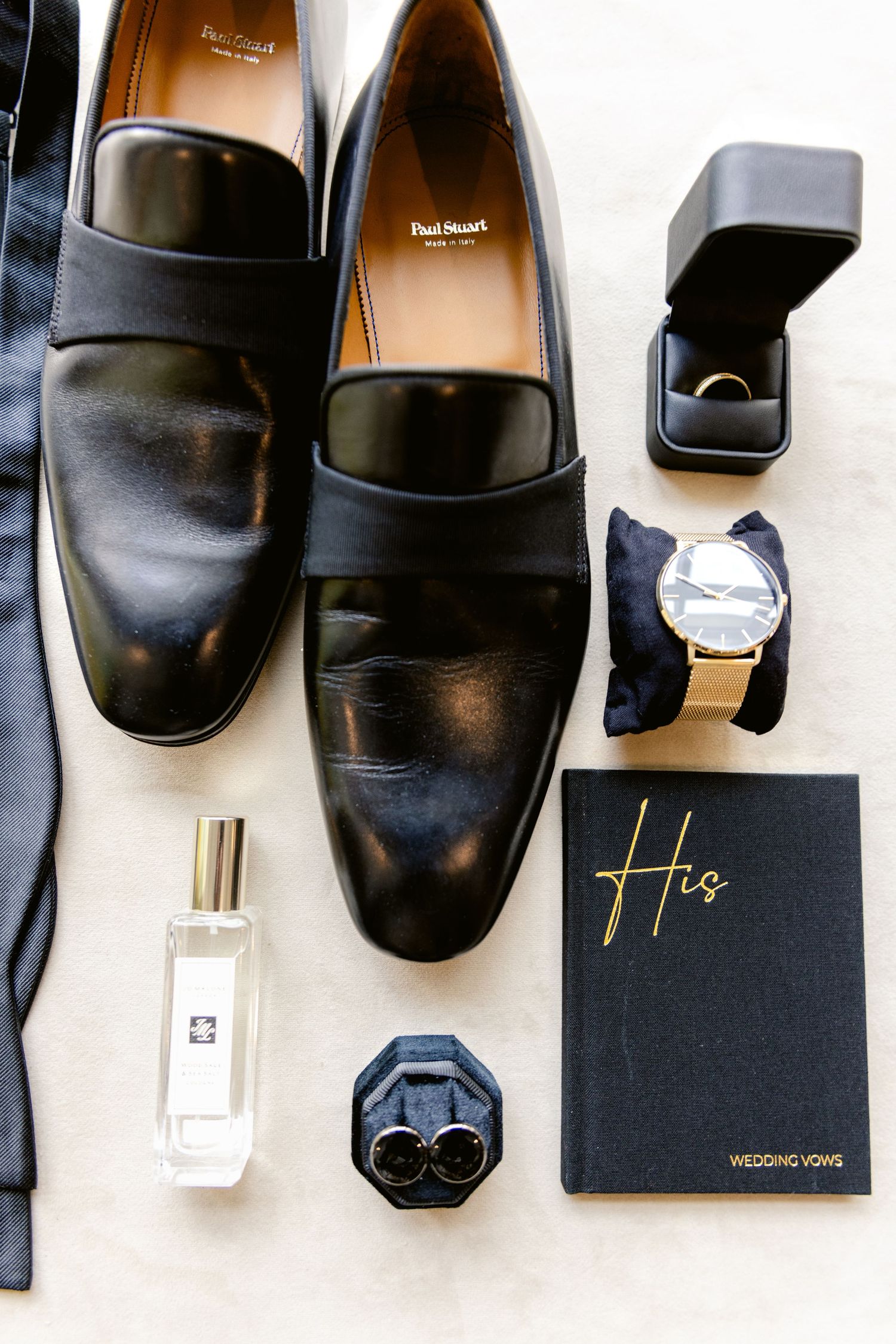 Black leather dress shoes and wedding accessories arranged on a white surface.
