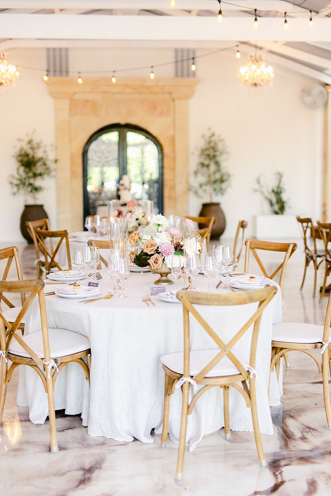 Elegant wedding reception venue with round tables, white linens, and wooden cross-back chairs in a bright airy space.