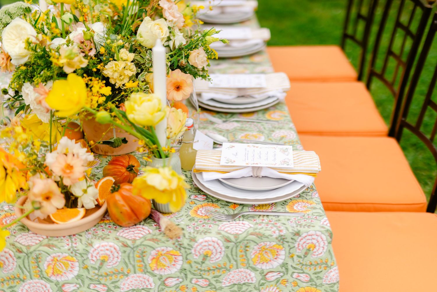A festive outdoor table setting with orange cushions, floral tablecloth, and yellow flower centerpiece arrangement.