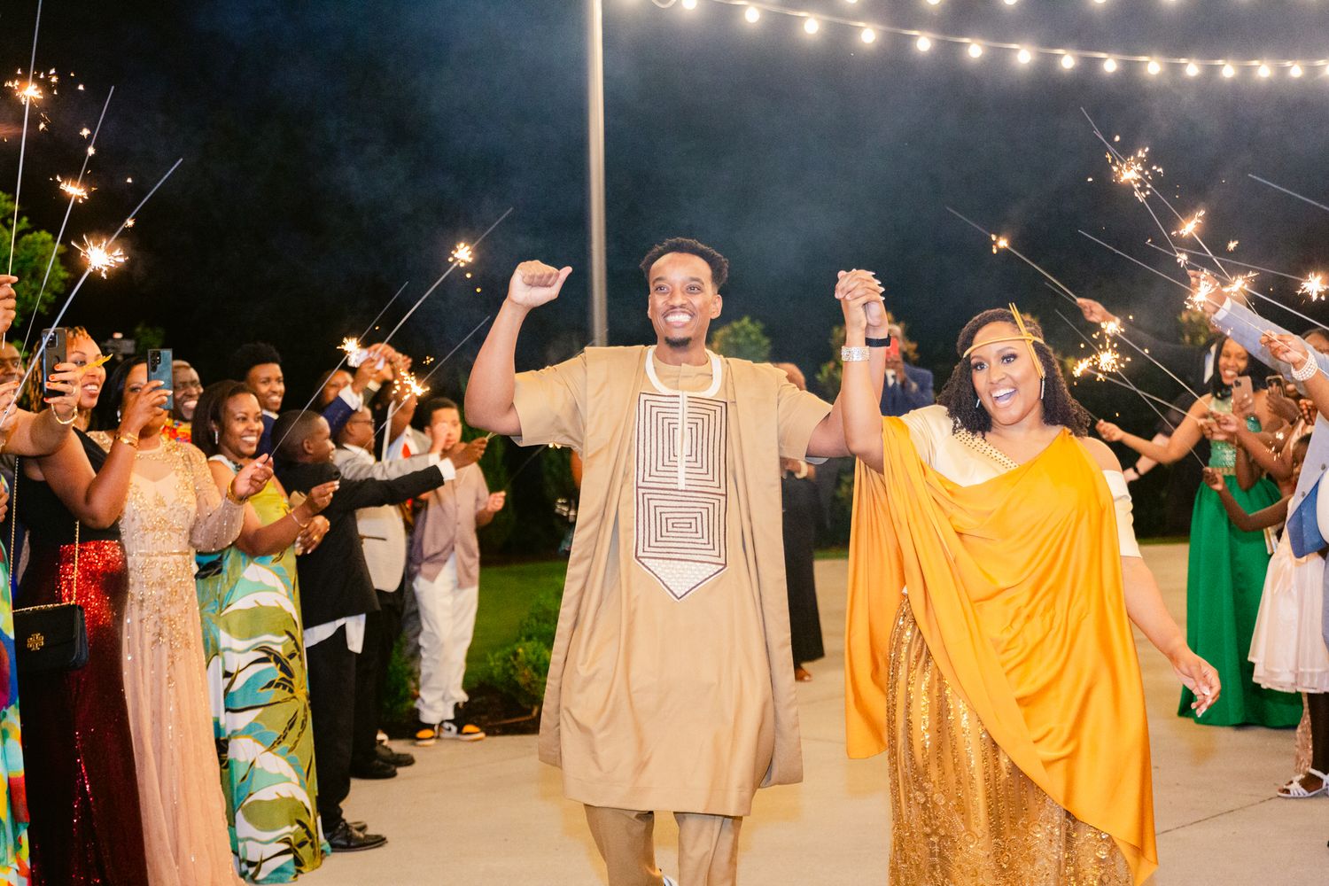 Wedding guests form a sparkler tunnel at night as couple walks through in traditional South Asian attire under string lights.