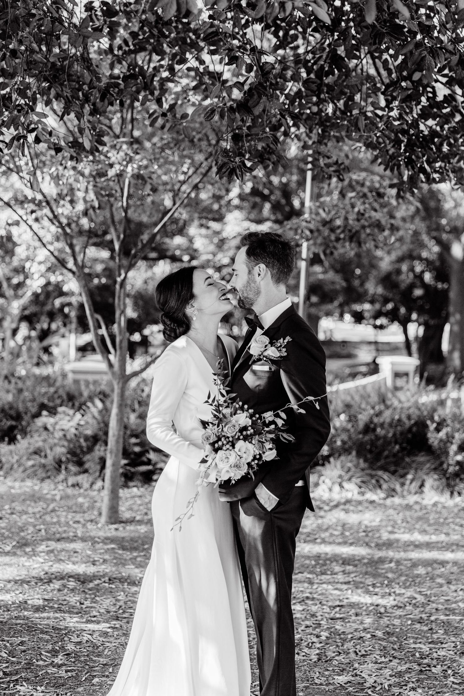 A romantic black and white wedding photo of a couple embracing under trees in a garden setting.